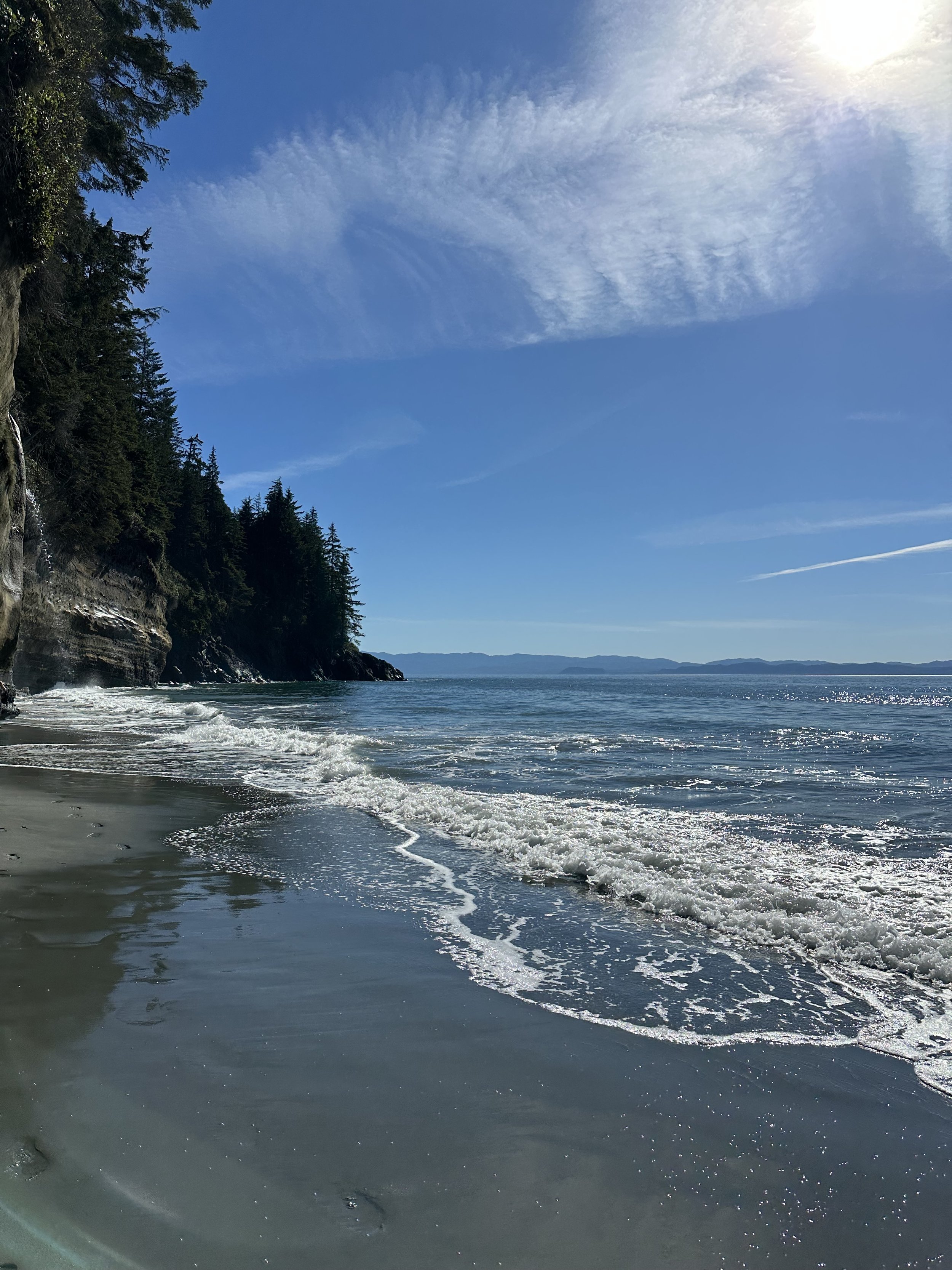 A scenic beach with waves gently crashing on the sandy shore, surrounded by forested cliffs to the left, under a partly cloudy blue sky.