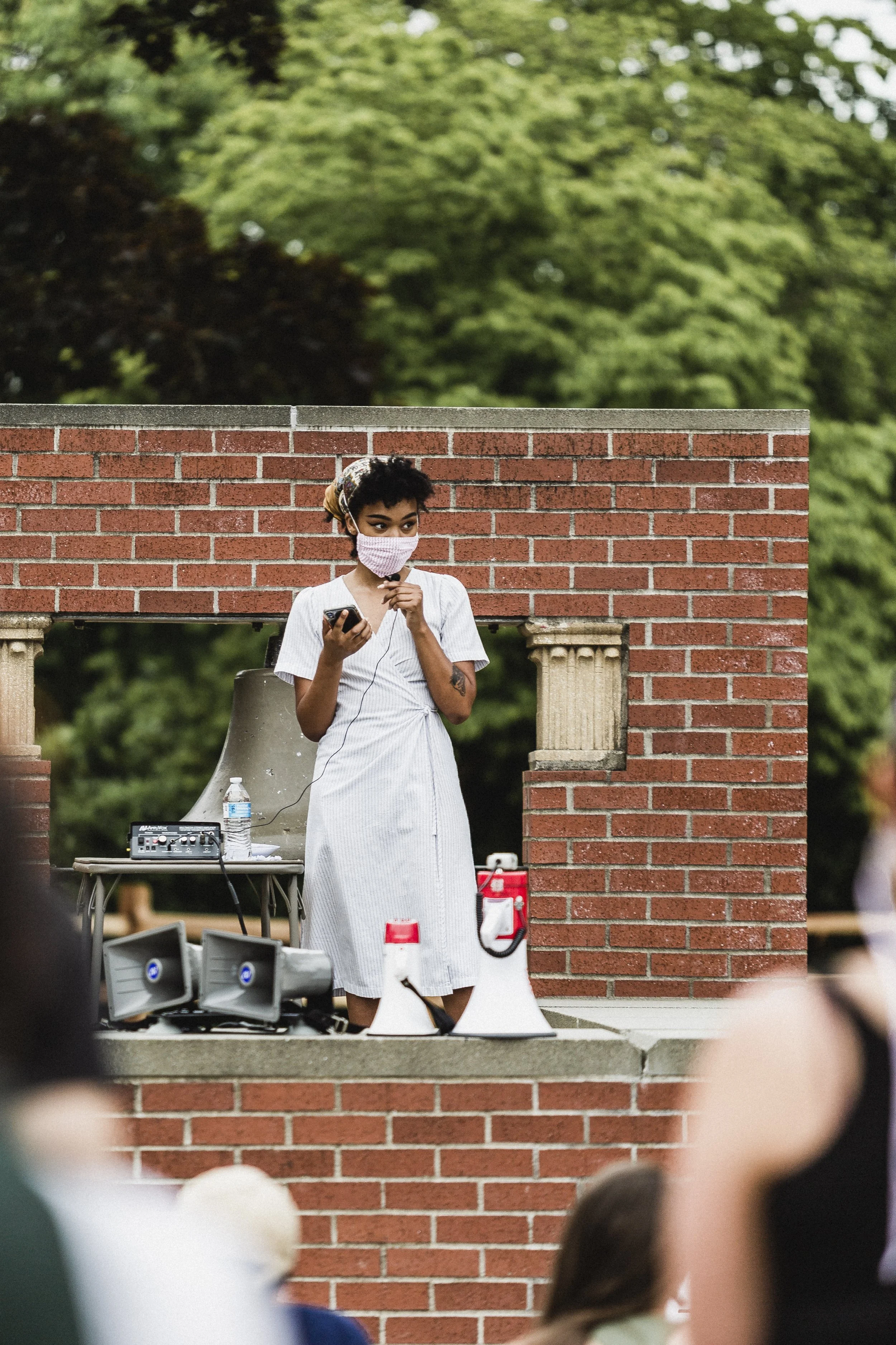 A woman wearing a pink face mask stands behind a brick wall, holding a microphone and looking at her phone. There are loudspeakers, a water bottle, and a fire extinguisher nearby.