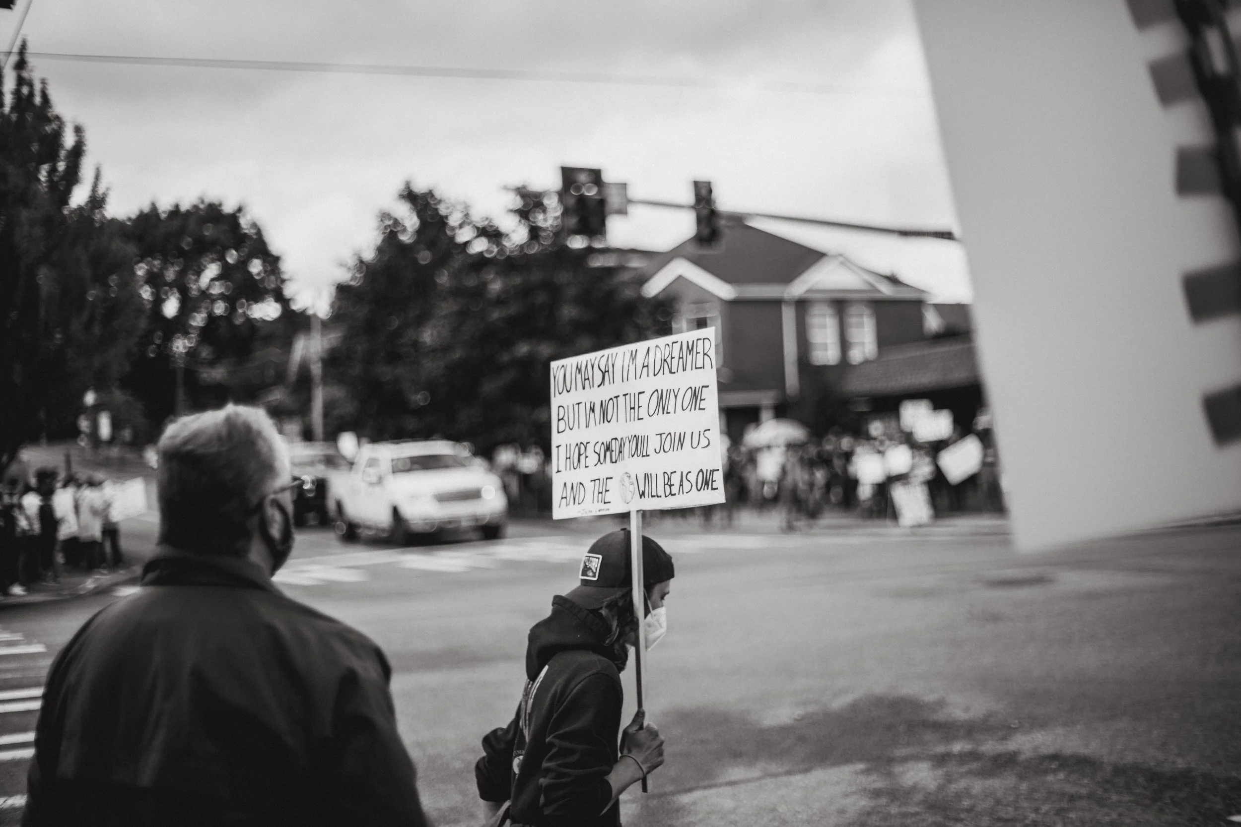Person holding a sign at a protest crossing street with cars and other protesters in the background, wearing a face mask and cap.