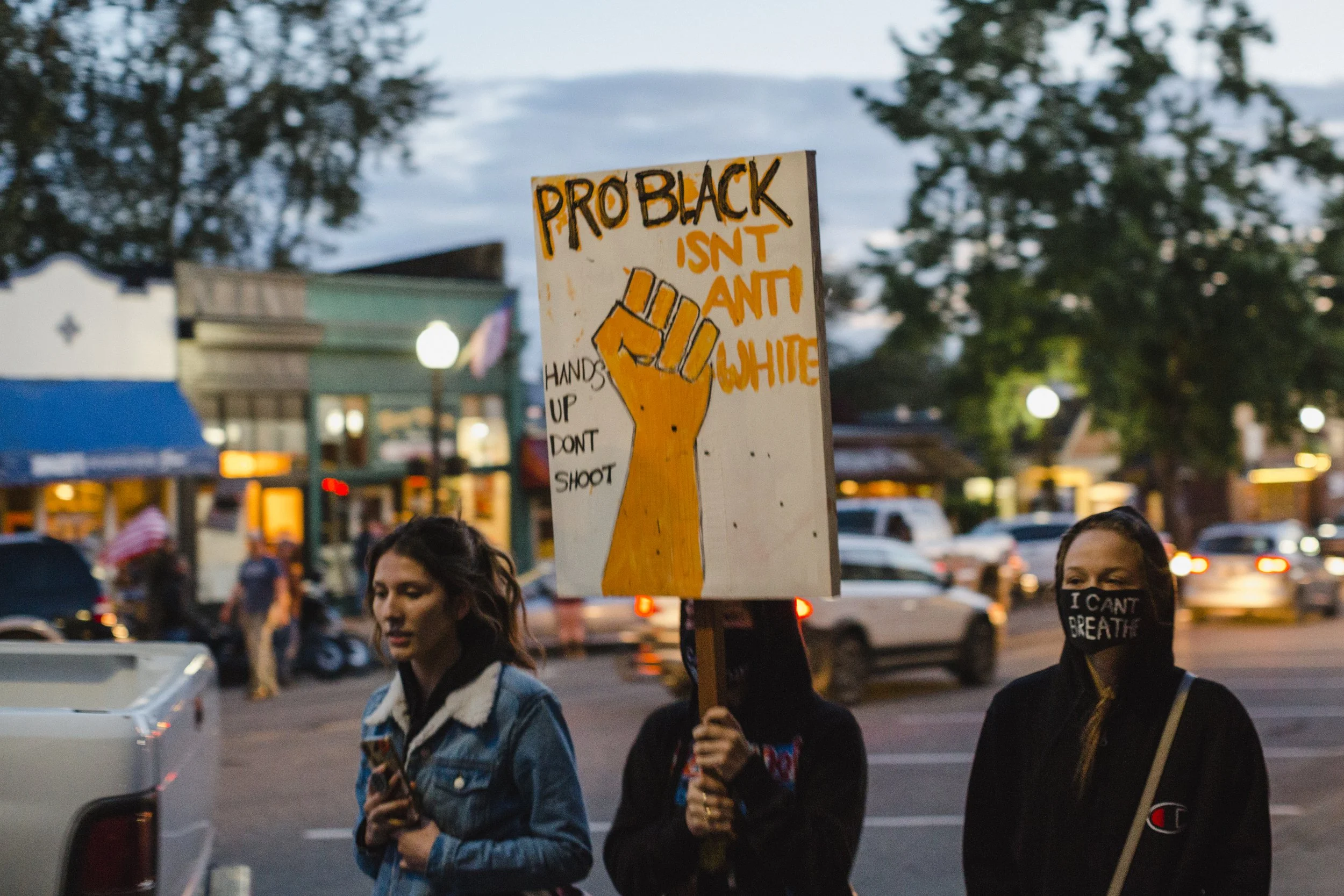 Protesters walking on a street holding signs with messages about racial justice, including one sign with a raised fist, that reads 'PRO BLACK ISN'T ANTI WHITE' and 'HANDS UP DON'T SHOOT'. One person is wearing a face mask that says 'I CAN'T BREATHE'.