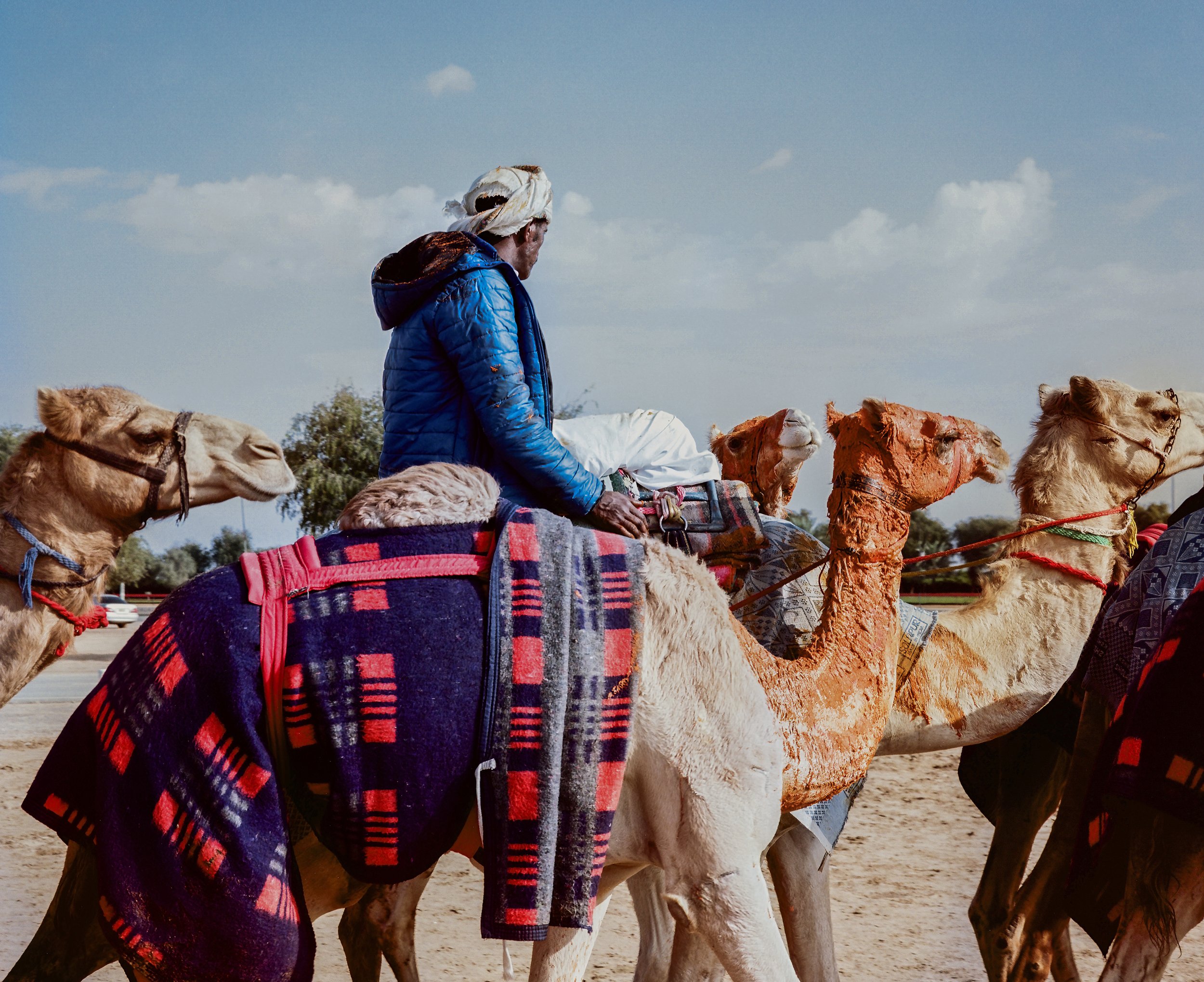 Man riding a camel with a blue jacket and white head covering in a desert setting, with others on camels behind him.