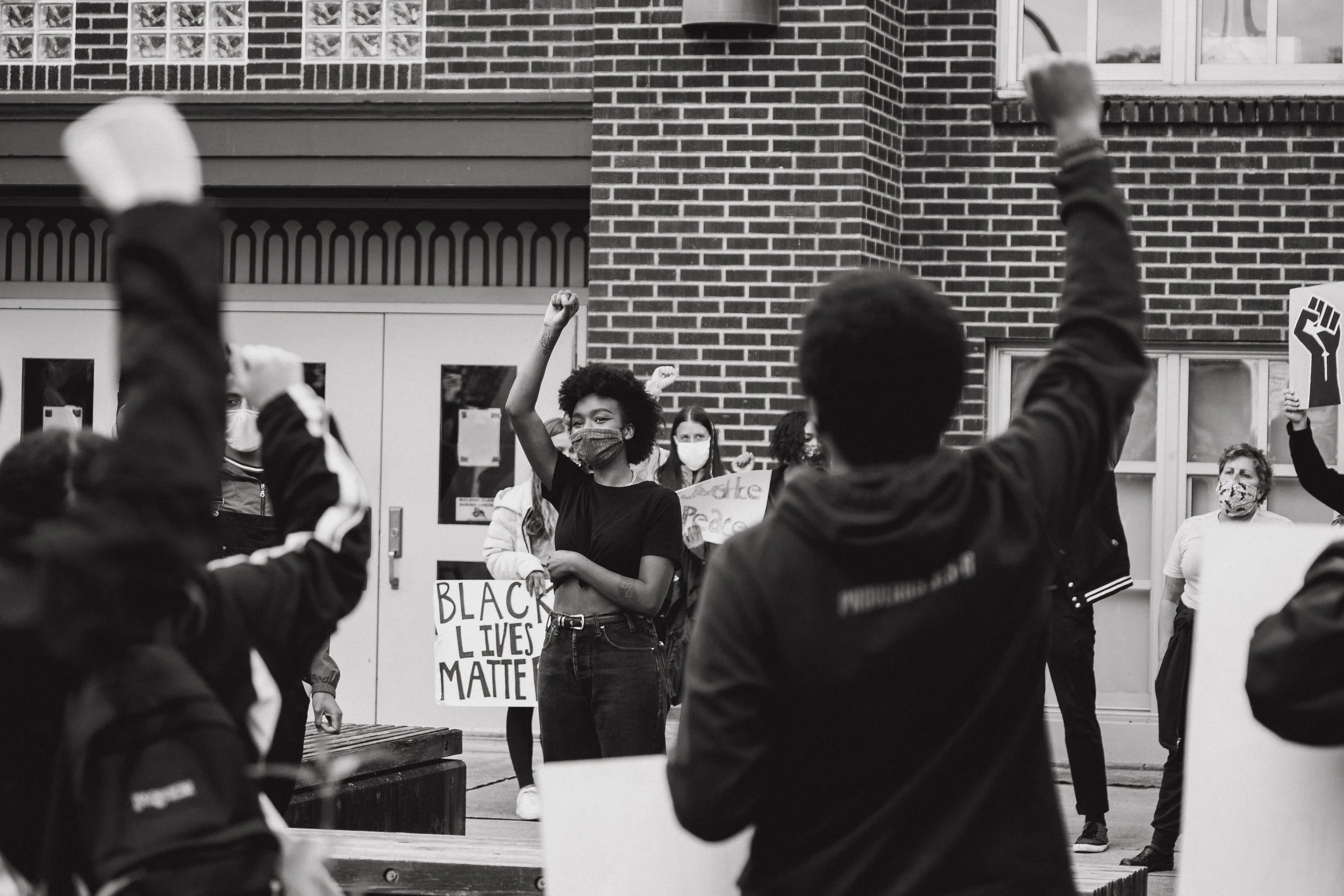 People protesting outside a brick building, holding signs supporting Black Lives Matter and equality, some wearing masks, raising fists.