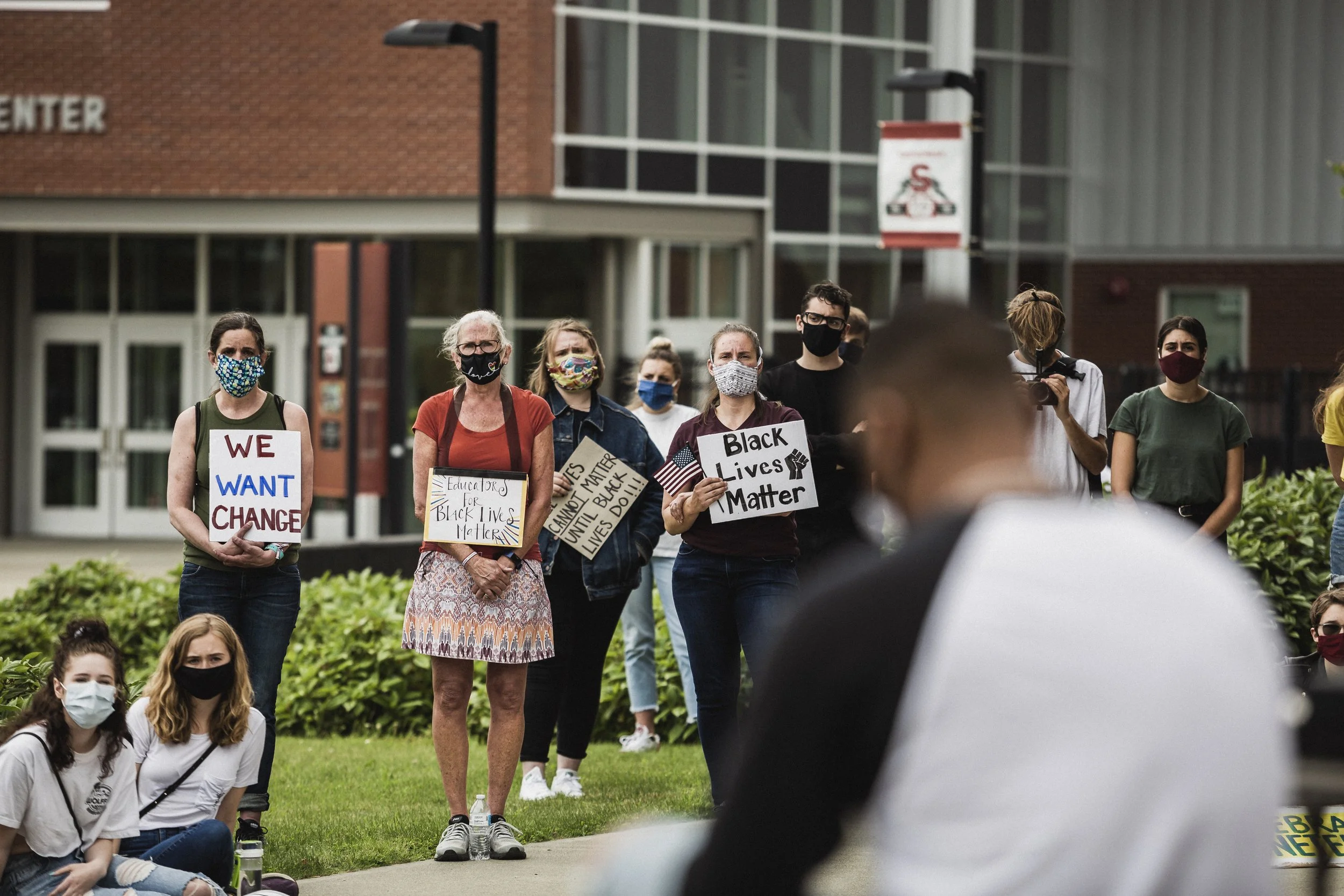Group of diverse people at a protest holding signs with messages like "We Want Change," "Black Lives Matter," and "Education for Black Lives Matter." Many are wearing masks.