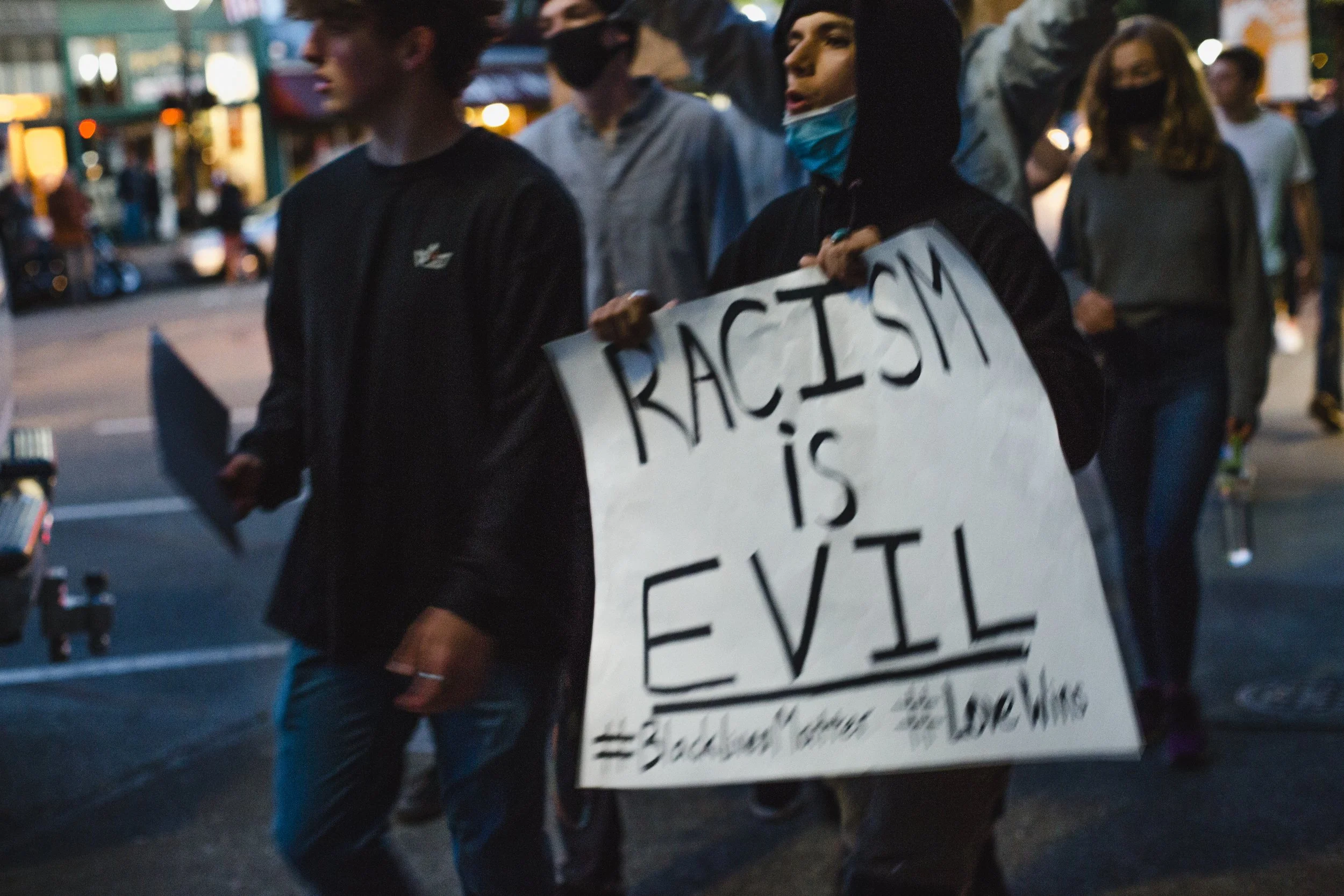 People participating in a protest march, with one person holding a sign that reads 'Racism is Evil', in an urban area during evening.