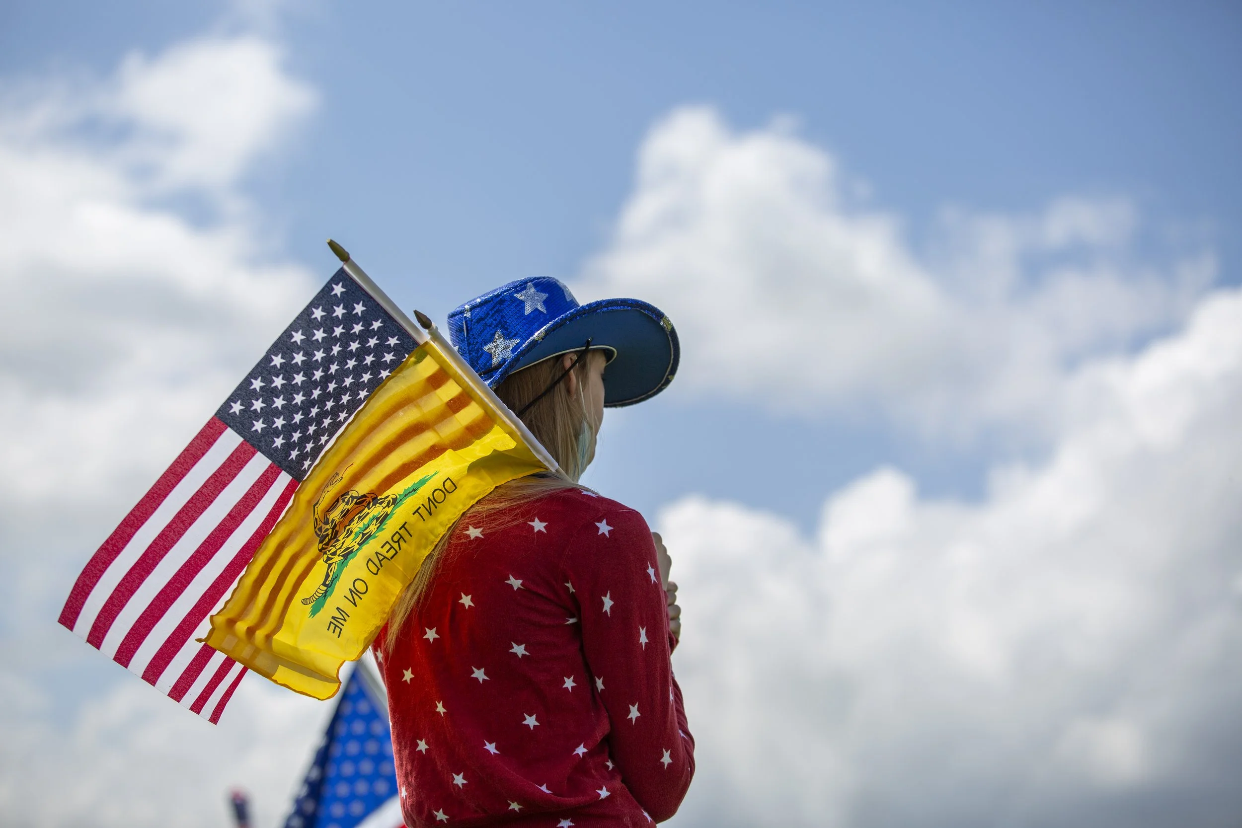 A woman wearing a red shirt with white stars, a blue cowboy hat with white stars, and a face mask, holding American and Gadsden flags against a cloudy sky background.