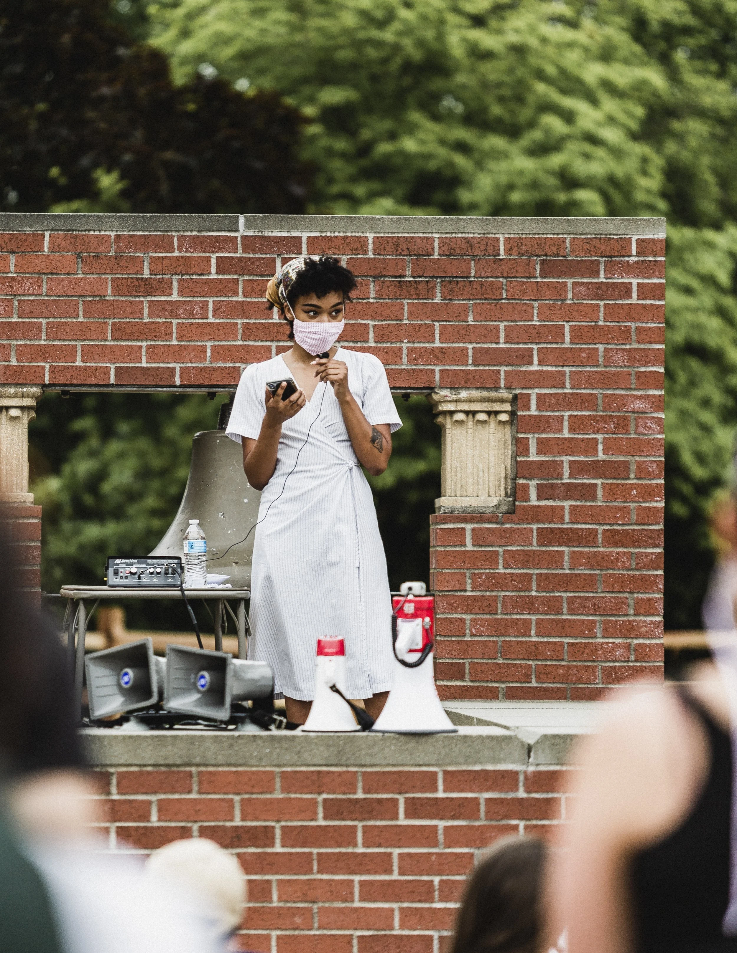 A woman with short curly hair wearing a face mask, white dress, and headphones stands behind a brick structure, holding a smartphone, with speakers, megaphones, and a water bottle on a small table nearby, outdoors in a park.