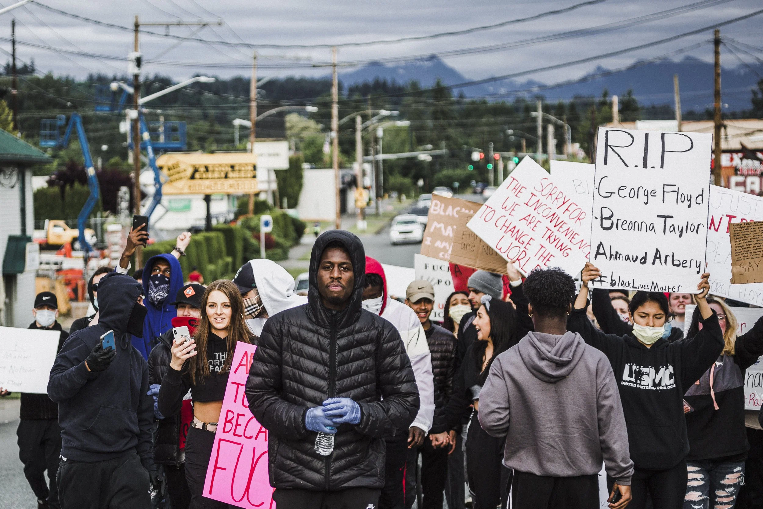 A diverse group of protesters holding signs and participating in a march on a street, with mountains and power lines in the background.