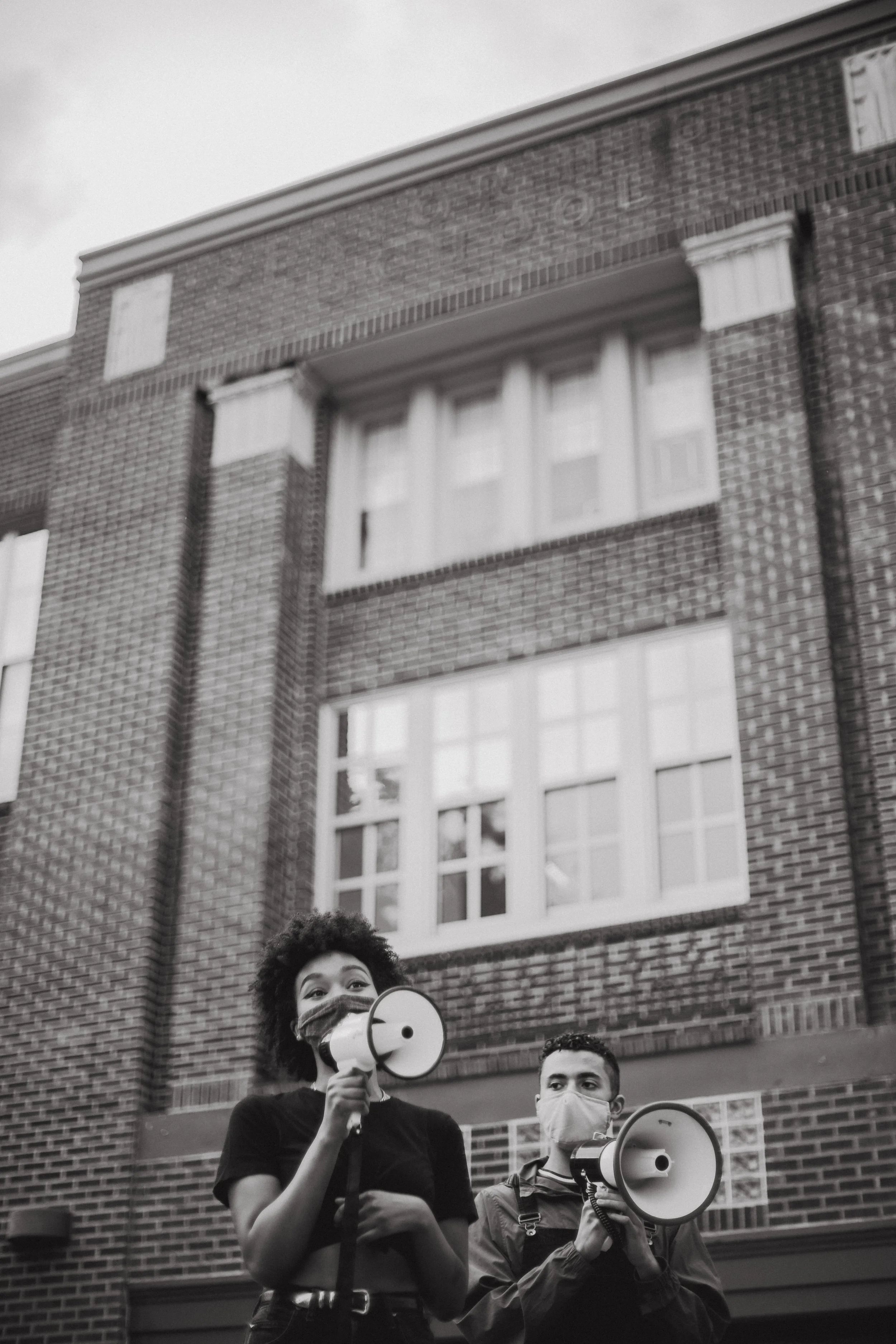 Two young people wearing masks holding megaphones protesting in front of a brick building.