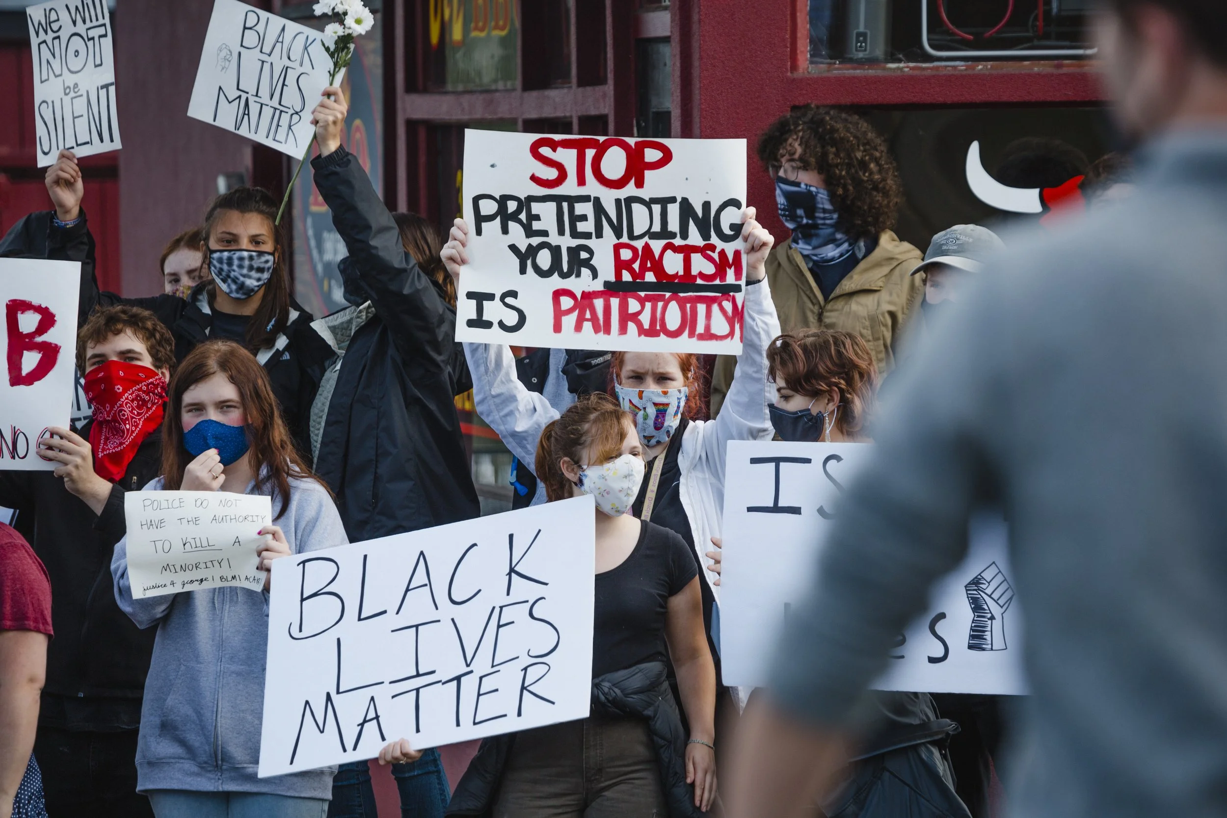 Group of protesters, mostly young, holding signs advocating for Black Lives Matter, with some signs reading "Black Lives Matter," "Stop pretending your racism is patriotism," and "We will not be silent." Many participants are wearing face masks.