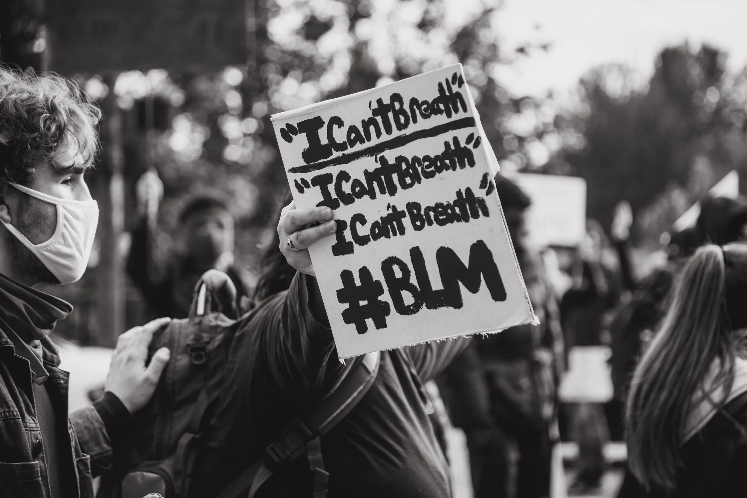 A person at a protest holding a sign that reads 'I Can't Breath' multiple times, with the hashtag #BLM at the bottom, while wearing a face mask.