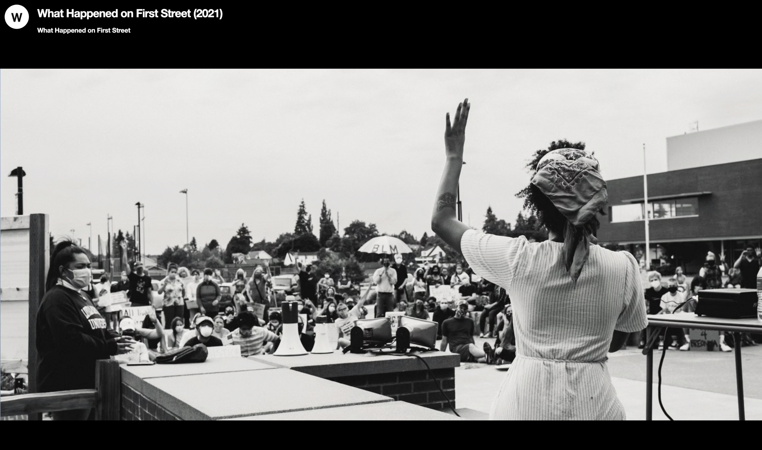 A Black woman with a bandana on her head raising her hand while speaking at a rally. Many protesters sit on the ground, some wearing masks, holding signs, and one umbrella with 'BLM' written on it. The scene is outdoors, with a modern building and trees in the background.