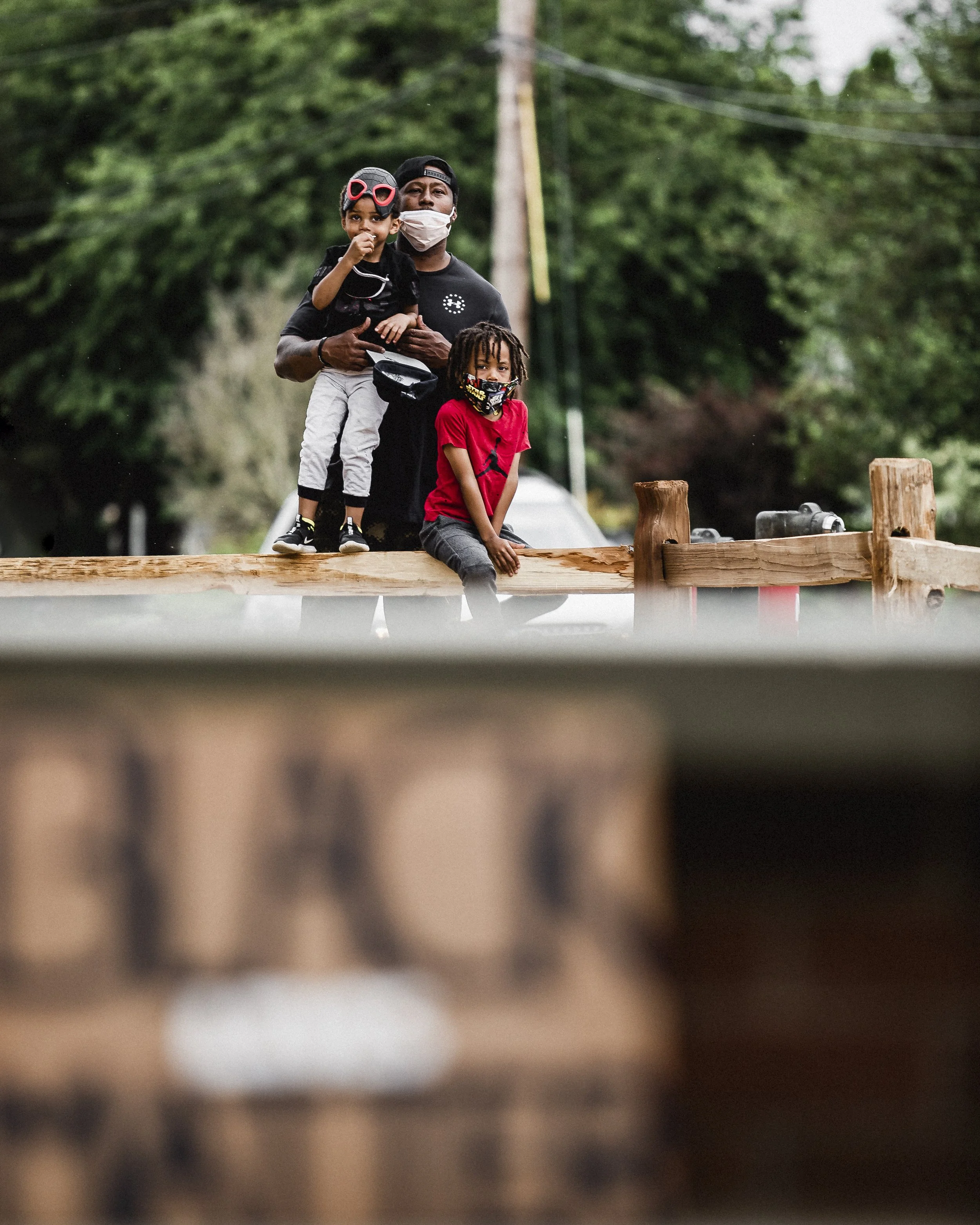 A man wearing a face mask holding a young girl, and a young boy sitting on a wooden railing outdoors with trees in the background.