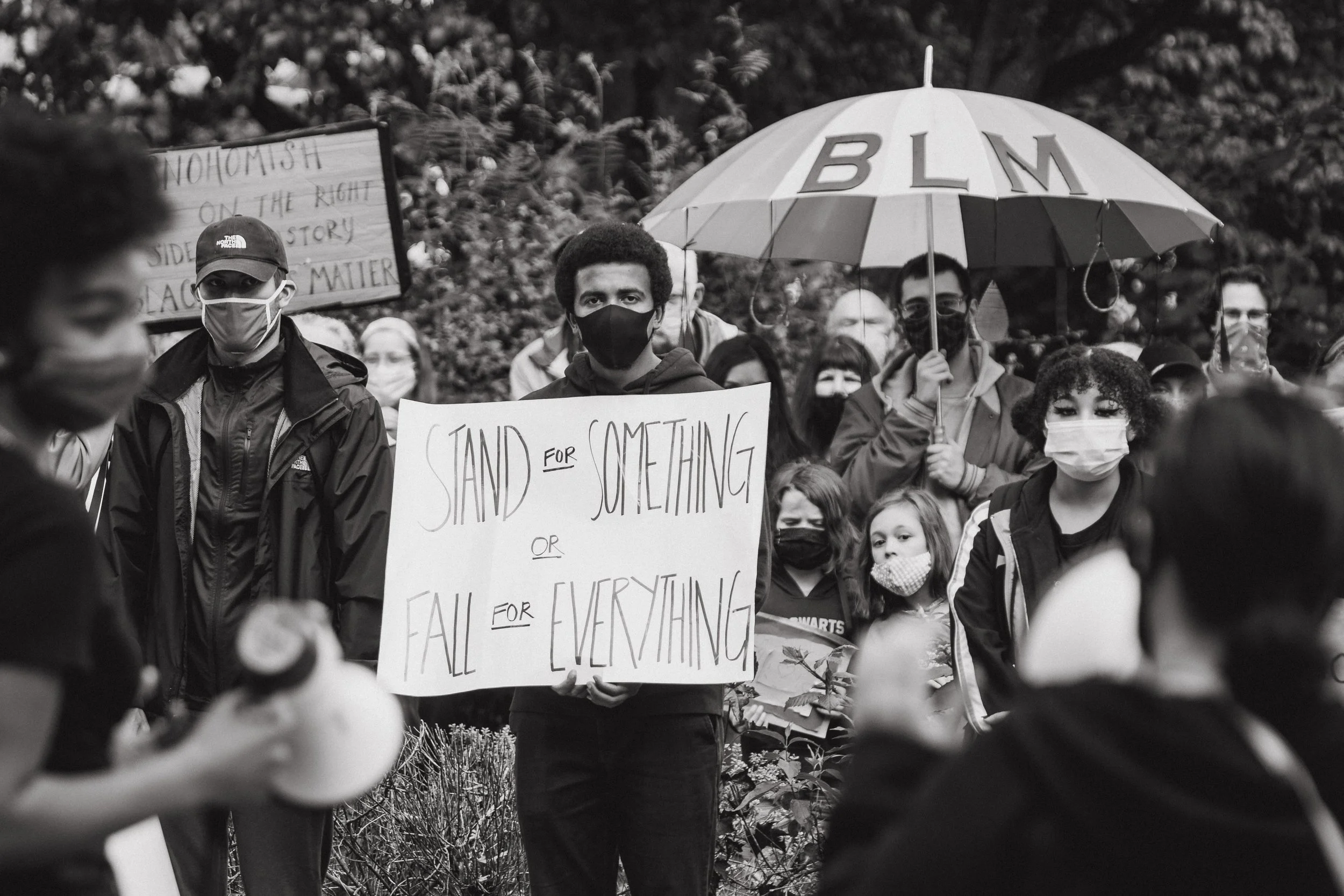 A group of diverse people gathered at a protest, many wearing masks. One person holds a sign that reads 'Stand for something or fall for everything,' while another holds an umbrella with the initials 'BLM' on it.