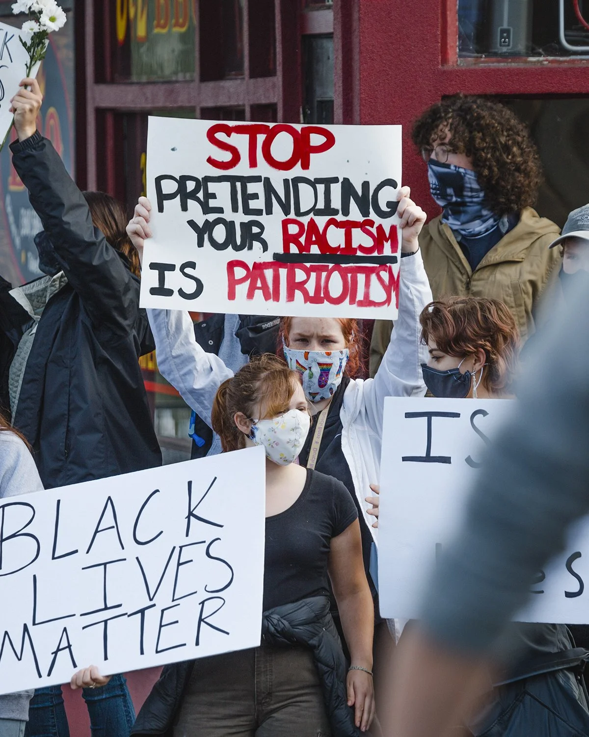 People participating in a protest holding signs that say 'Stop pretending your racism is patriotism' and 'Black Lives Matter'.