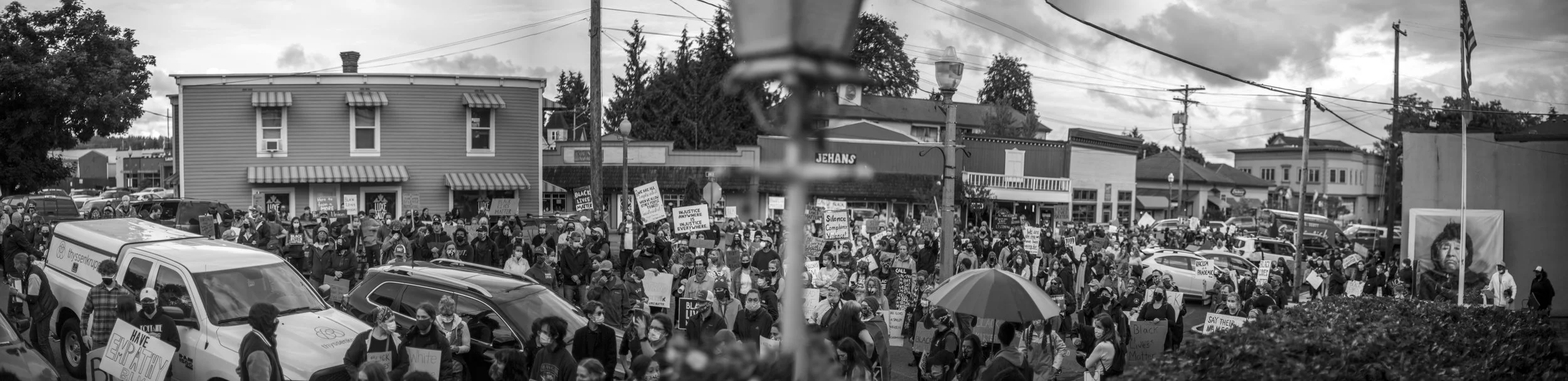 Black and white photo of a large protest with many people holding signs, on a street lined with shops and parked cars, some wearing masks, with a cloudy sky overhead.