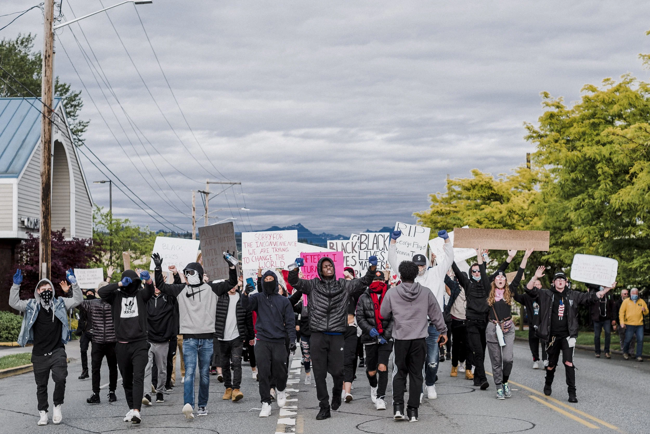 A large group of protesters marching down a street holding signs and raising their fists. The signs include messages such as 'Black Lives Matter', 'Sorry for the inconvenience, we are trying to change the world,' and 'R.I.P. George Floyd'. The group 