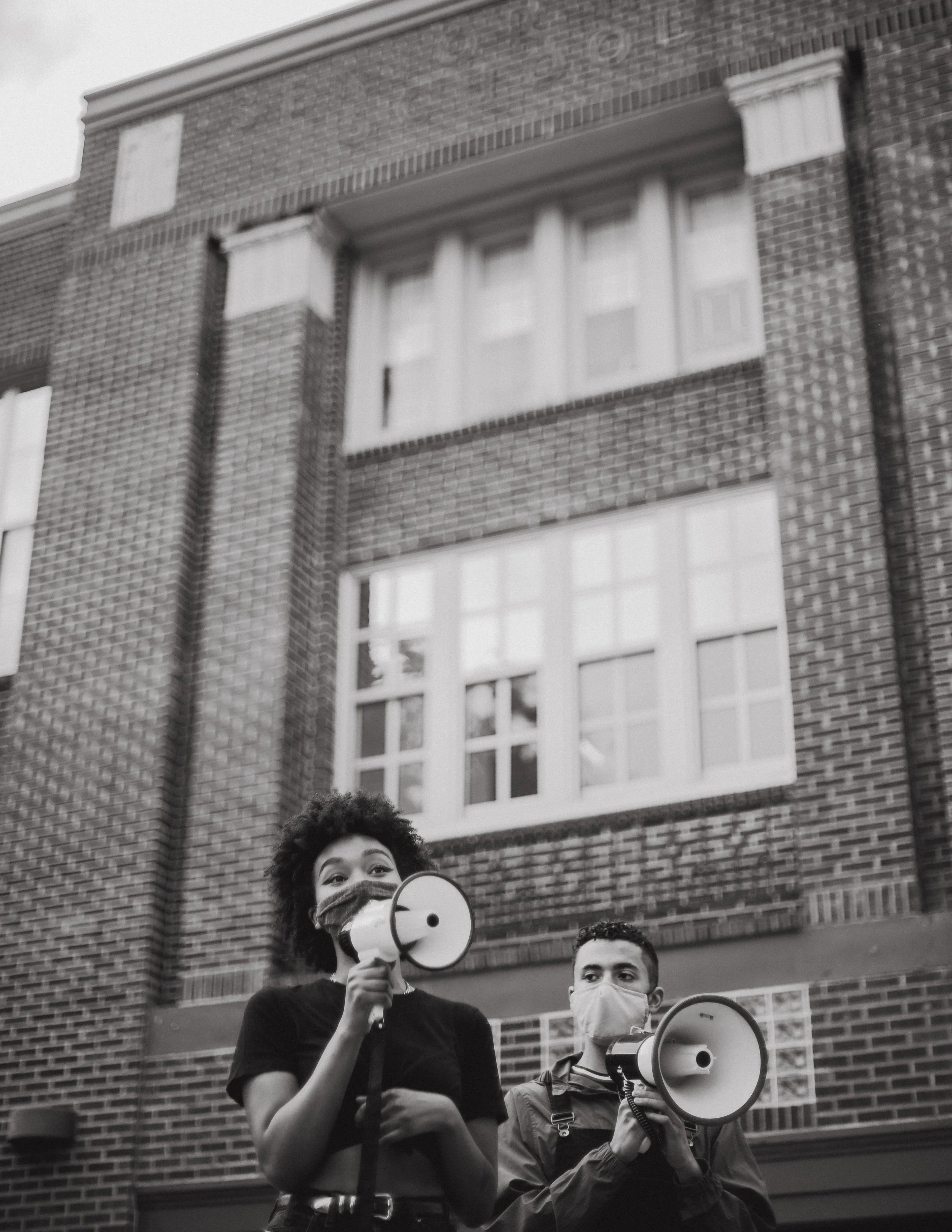 Black and white photo of two young people wearing masks, standing in front of a brick building, holding megaphones.