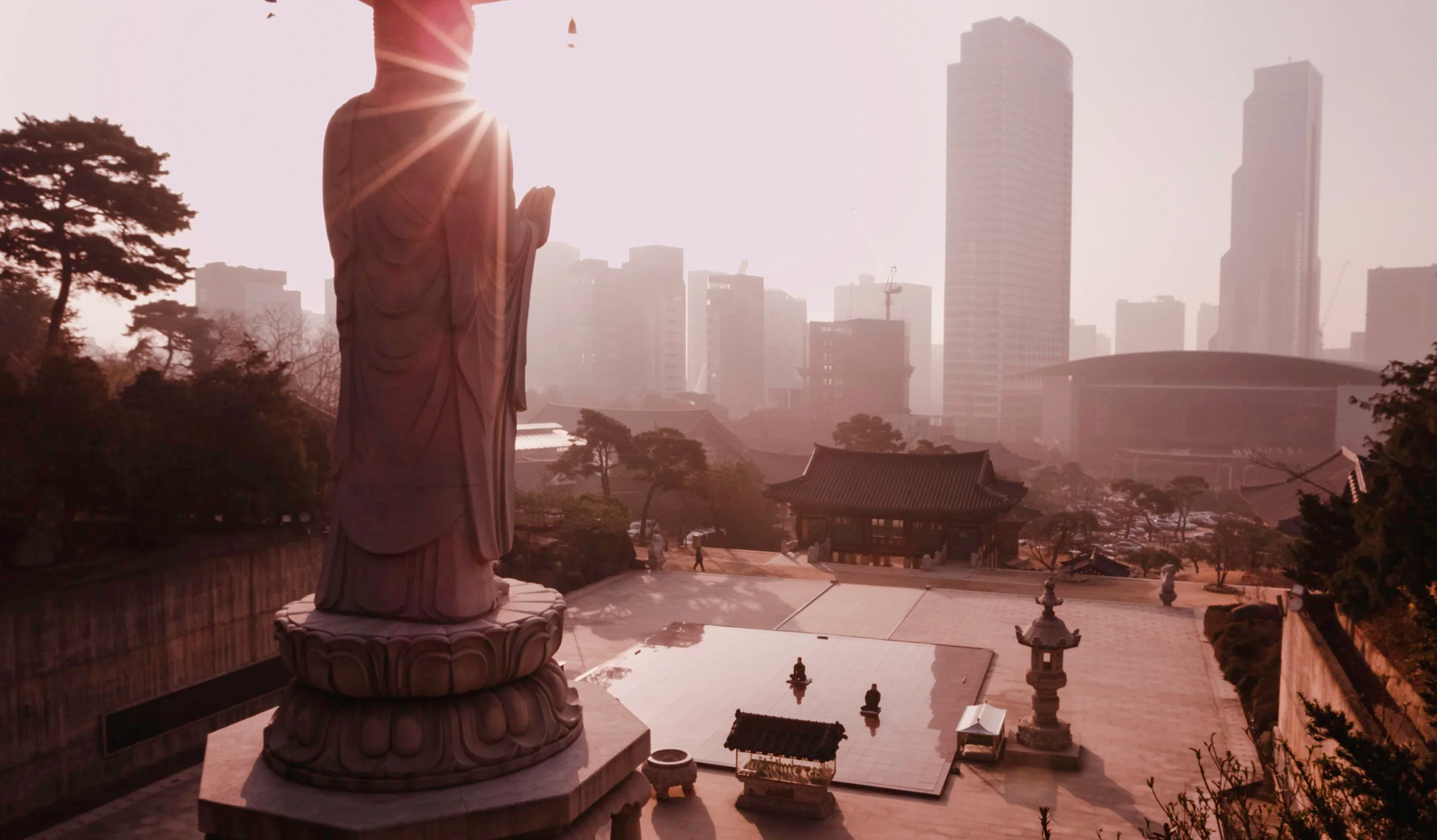 A large Buddha statue in a traditional pose is in the foreground, with the sun behind its head creating a starburst effect. Behind it, there are trees, a traditional Japanese-style building, and a city skyline with modern skyscrapers under a hazy sky.