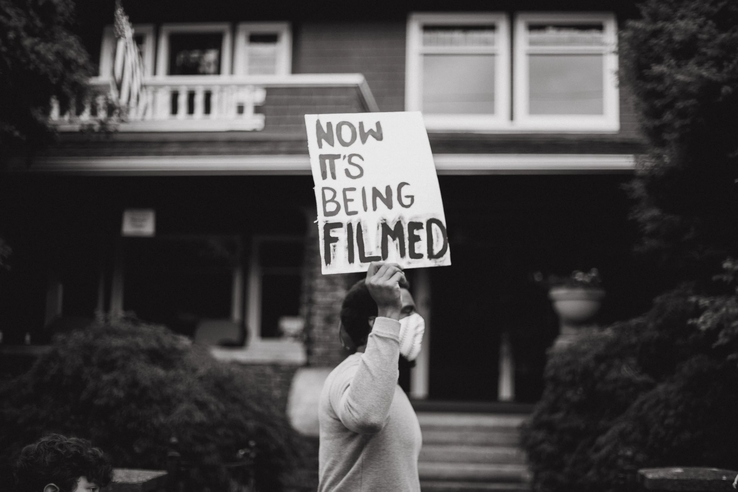 A person wearing a mask holding a sign that says "Now it's being filmed" in front of a house.
