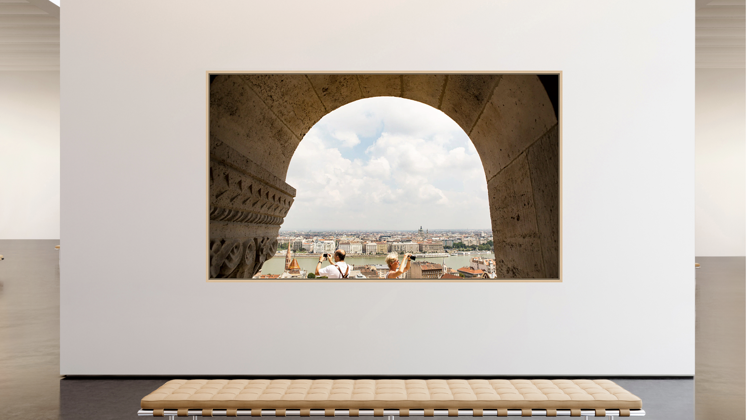 View through a large, arched window showing two tourists taking photos of a cityscape with a river, buildings, and a cloudy sky in Budapest.