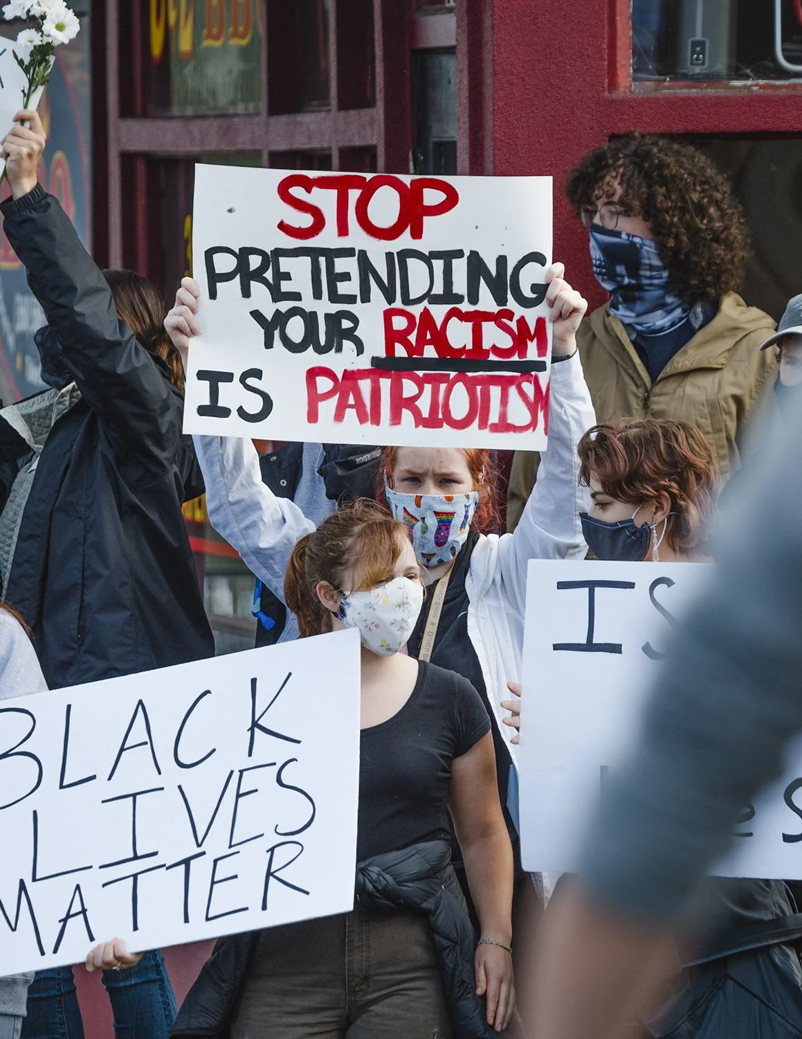 People protesting with signs, including one that says "Stop pretending your racism is patriotism," and another that says "Black Lives Matter." They are wearing masks.