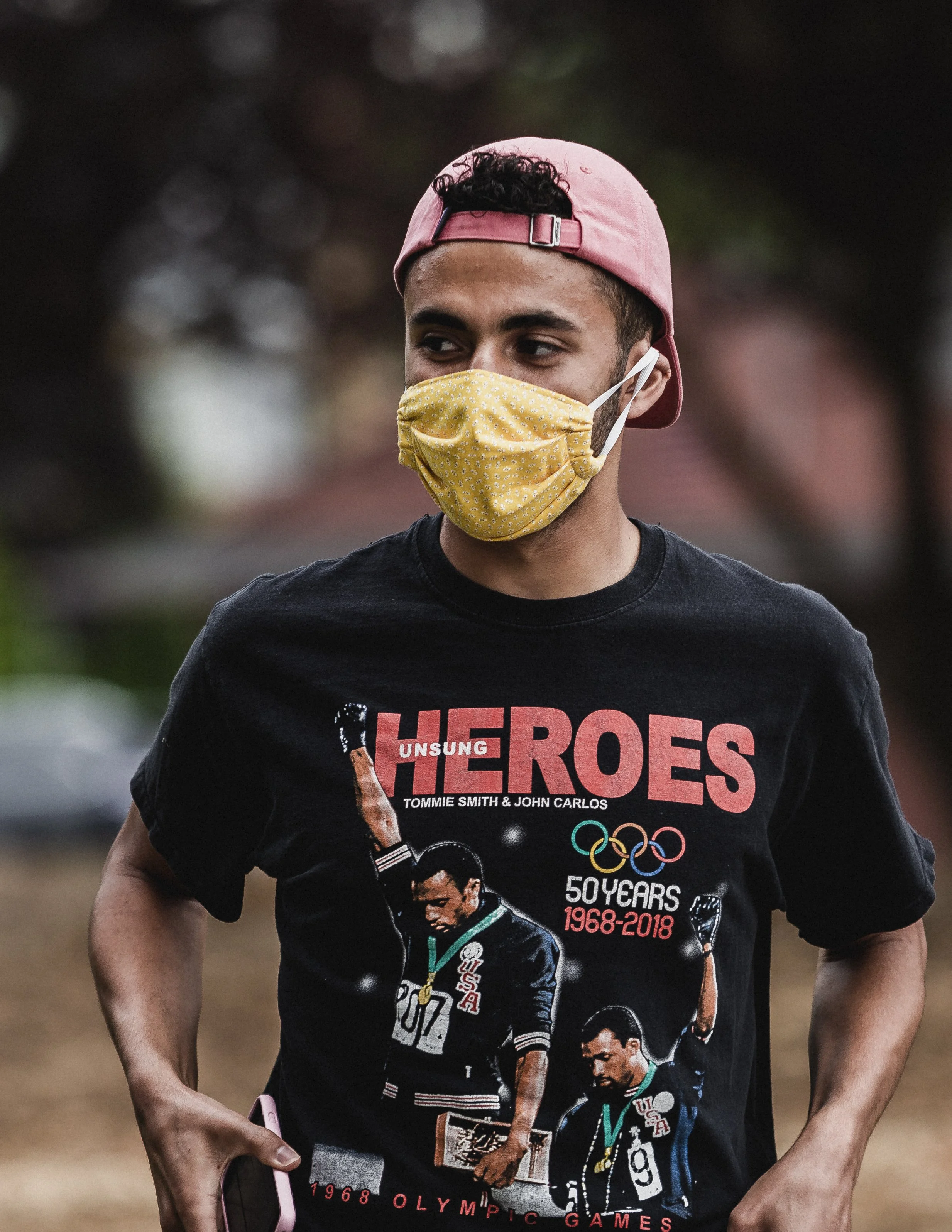 A young man wearing a pink baseball cap backwards and a yellow face mask. He is dressed in a T-shirt commemorating the 50th anniversary of the 1968 Olympics, featuring an image of an athlete with medals.