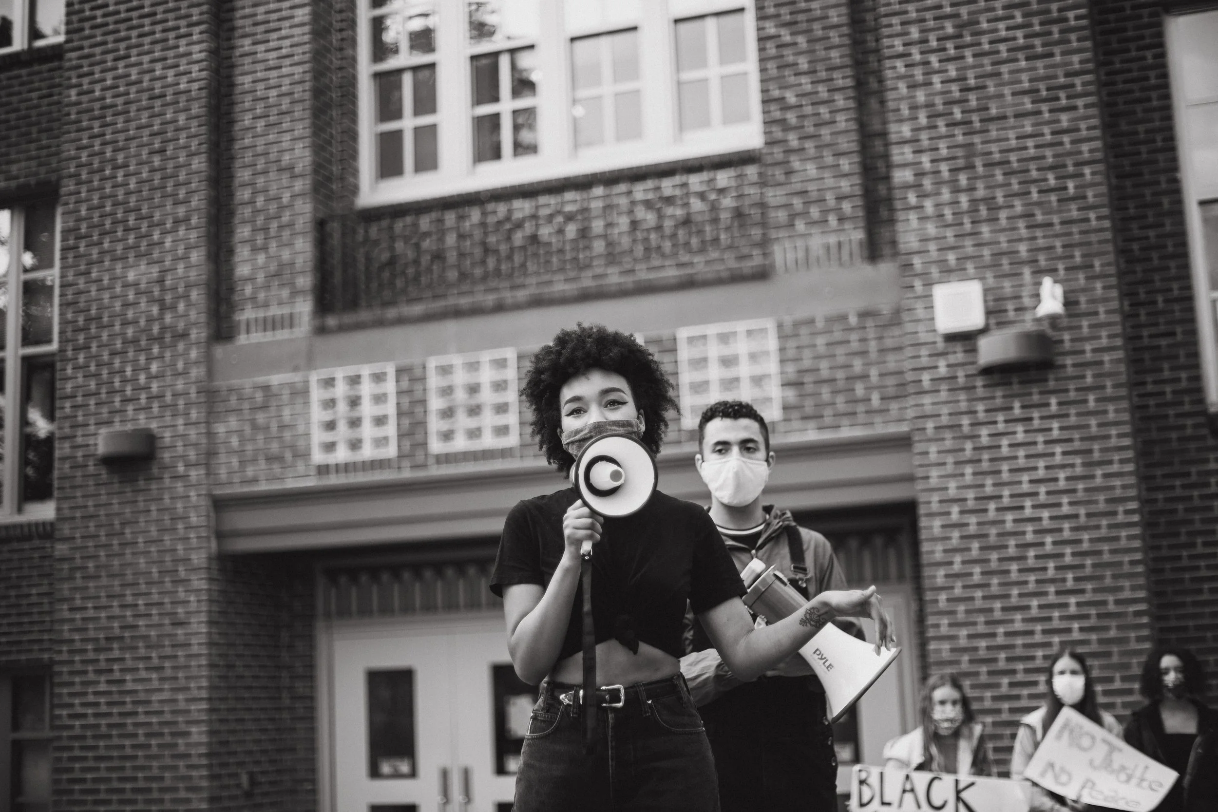 A woman with curly hair holding a megaphone and speaking, standing in front of a brick building, with people holding protest signs in the background, all wearing masks.