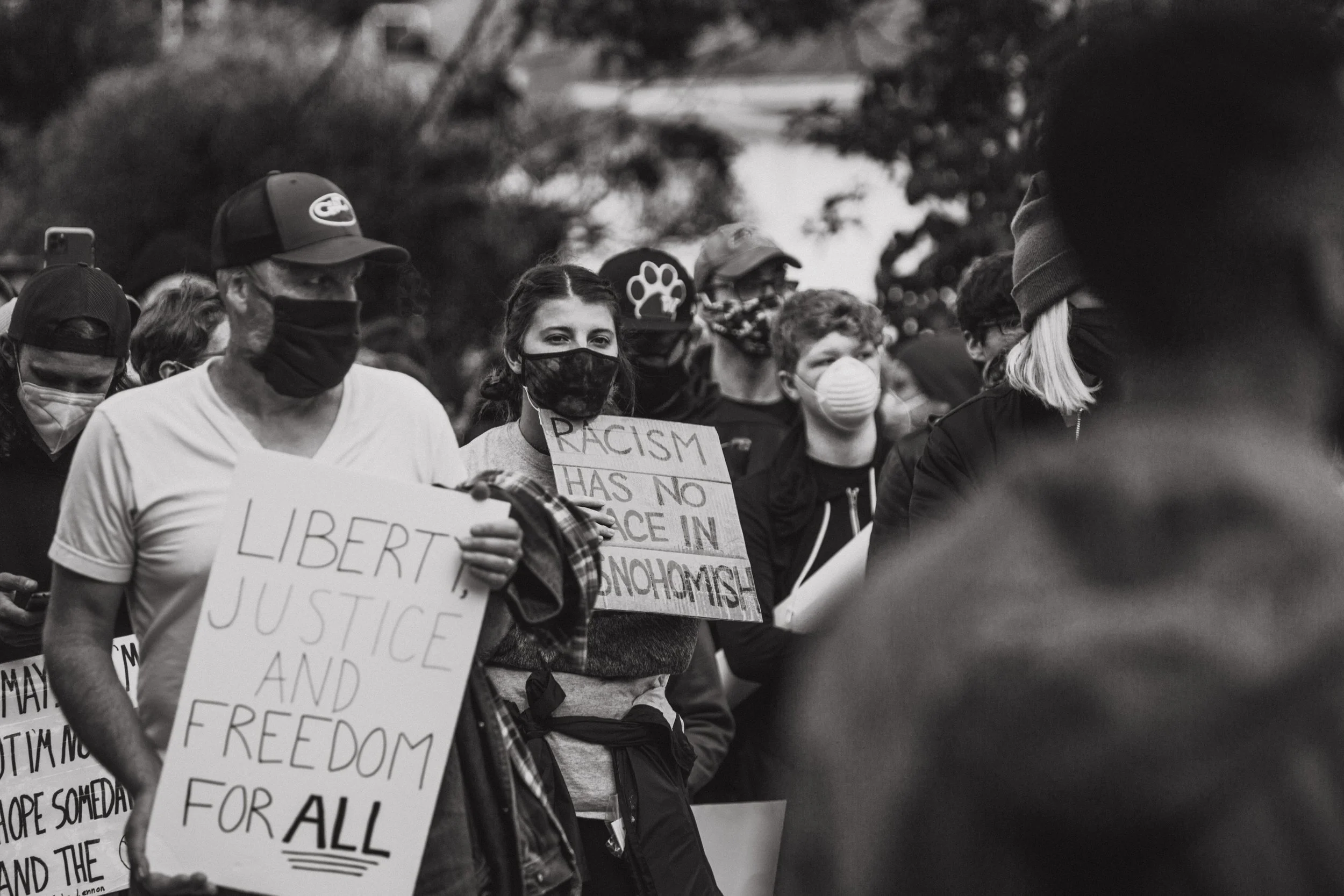 Group of people wearing masks at a protest, holding signs with messages such as "Liberty, Justice and Freedom for All" and "Racism Has No Place in Mononism."