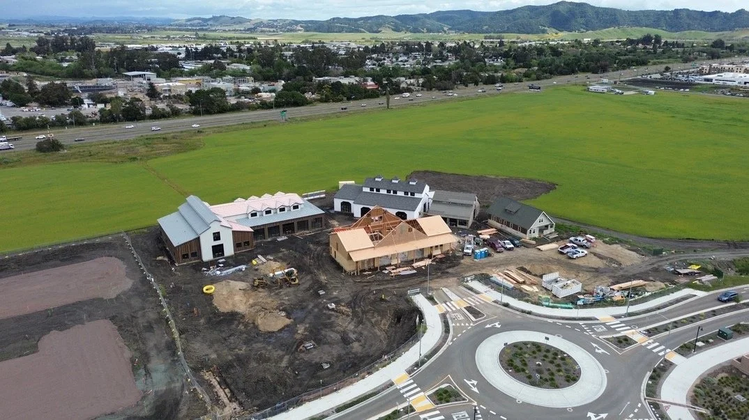 Aerial view of a construction site with multiple buildings in progress, surrounded by green fields and a roundabout in the foreground.