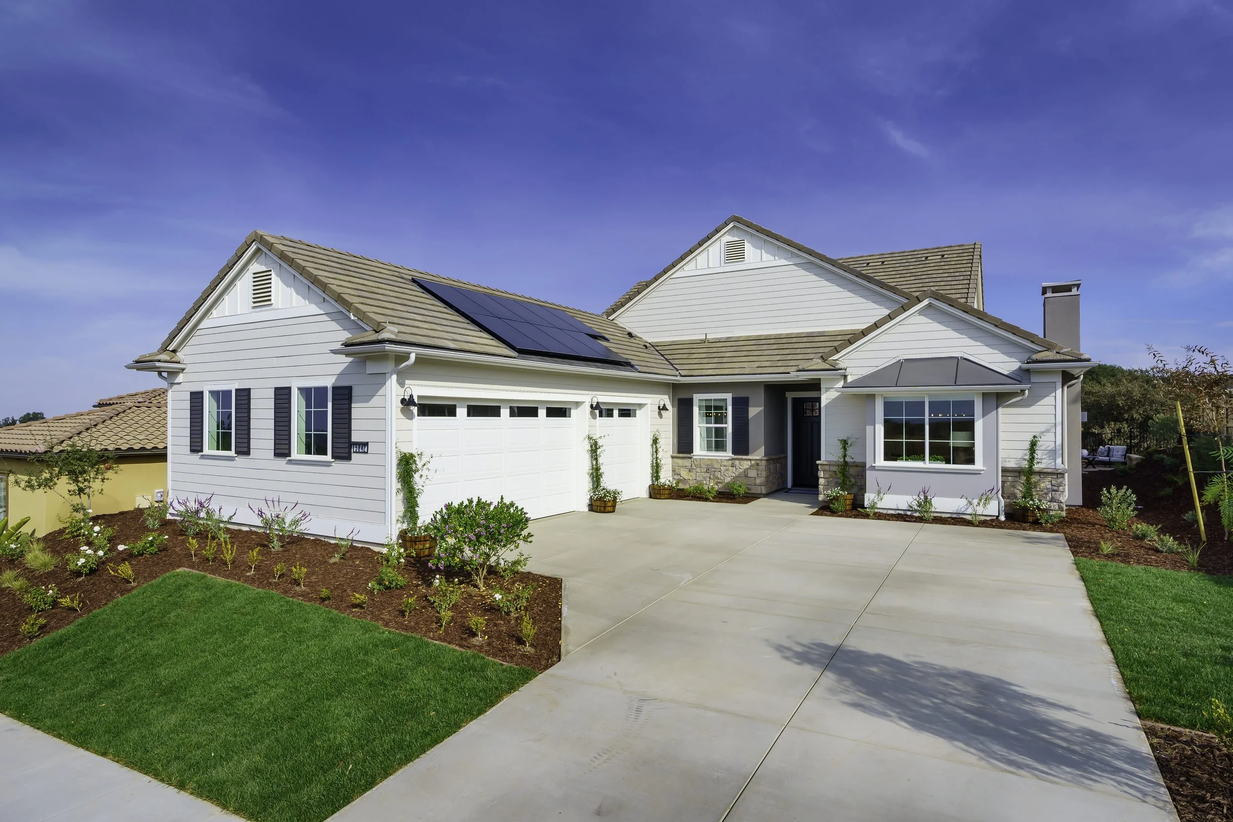 White house with a gray roof and solar panels, surrounded by a freshly landscaped yard with a concrete driveway and garden beds, under a blue sky.