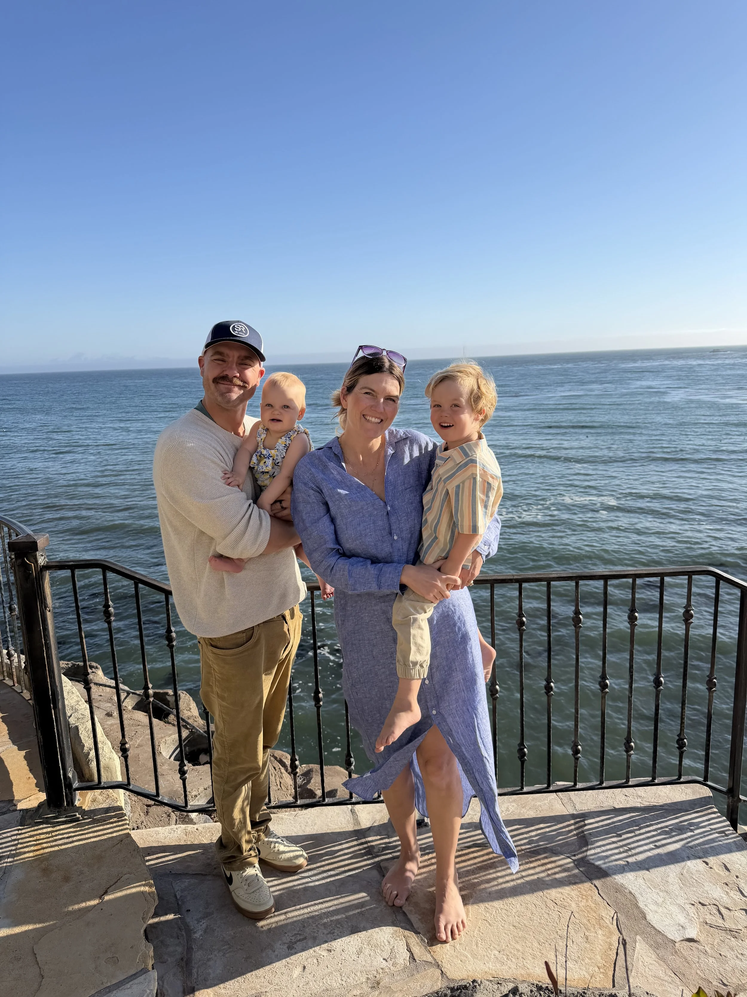 A family of four standing by the ocean on a sunny day, with a rocky walkway and metal railing in the foreground. Two adults and two children are smiling at the camera, with the ocean and a clear blue sky behind them.