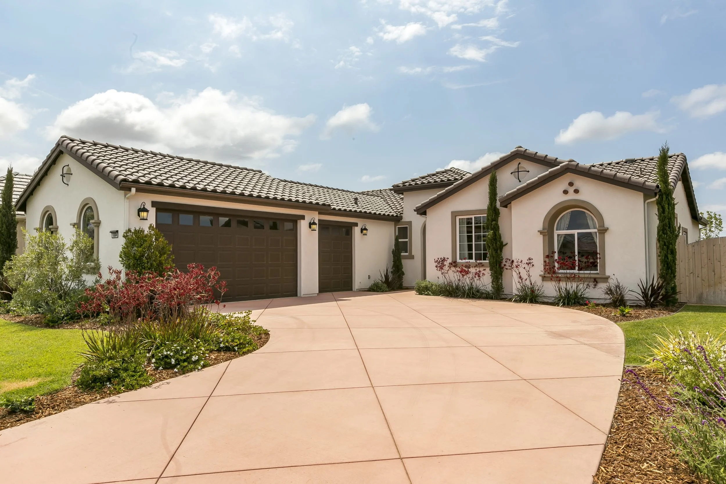 A modern house with a tiled roof, two garage doors, and a landscaped front yard with bushes, small trees, and flowering plants. The driveway is light-colored concrete, and the sky is partly cloudy.