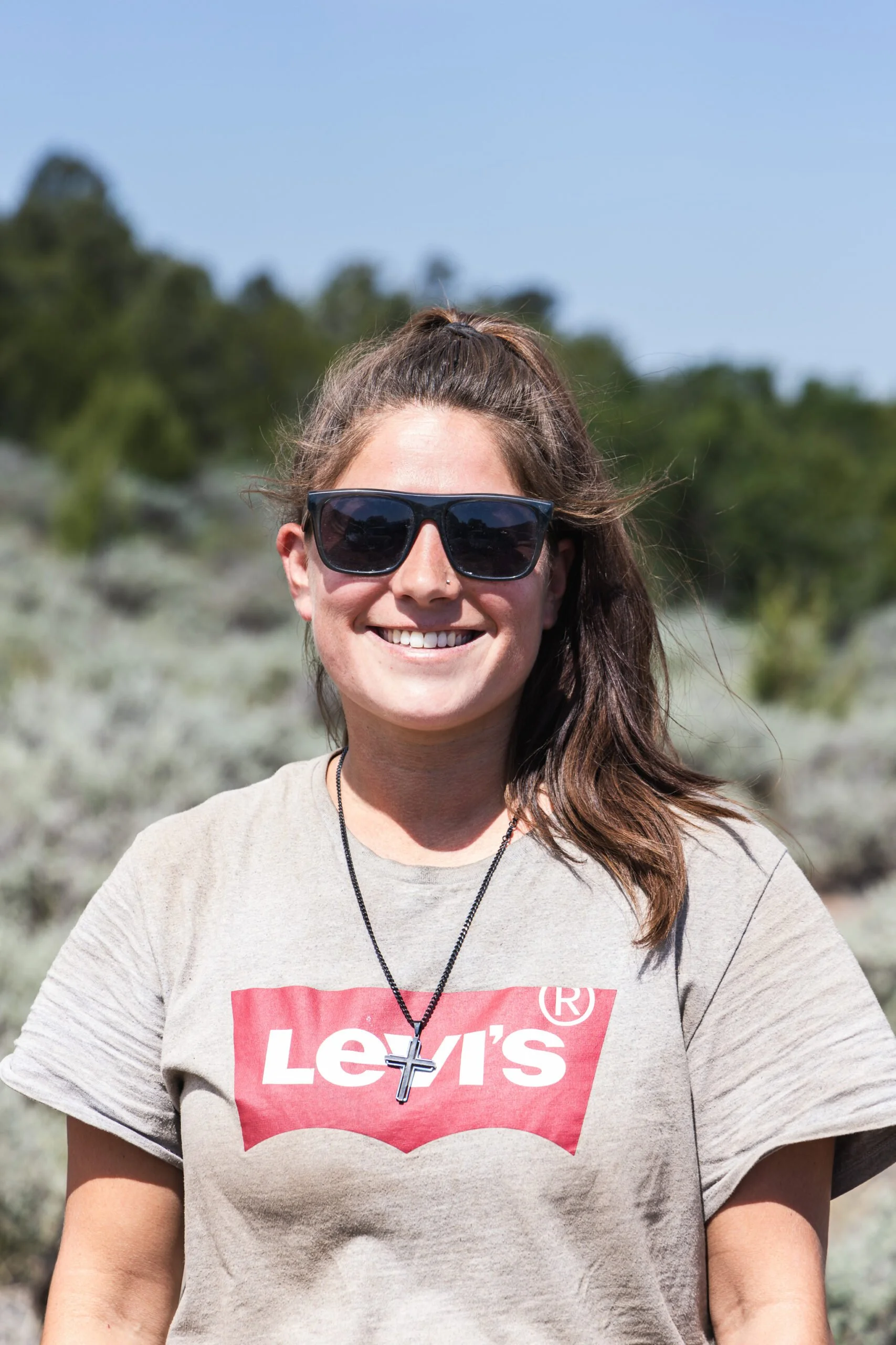 A smiling woman with long brown hair, wearing black sunglasses, a gray Levi's t-shirt, a cross necklace, standing outdoors with a blurred natural landscape background.