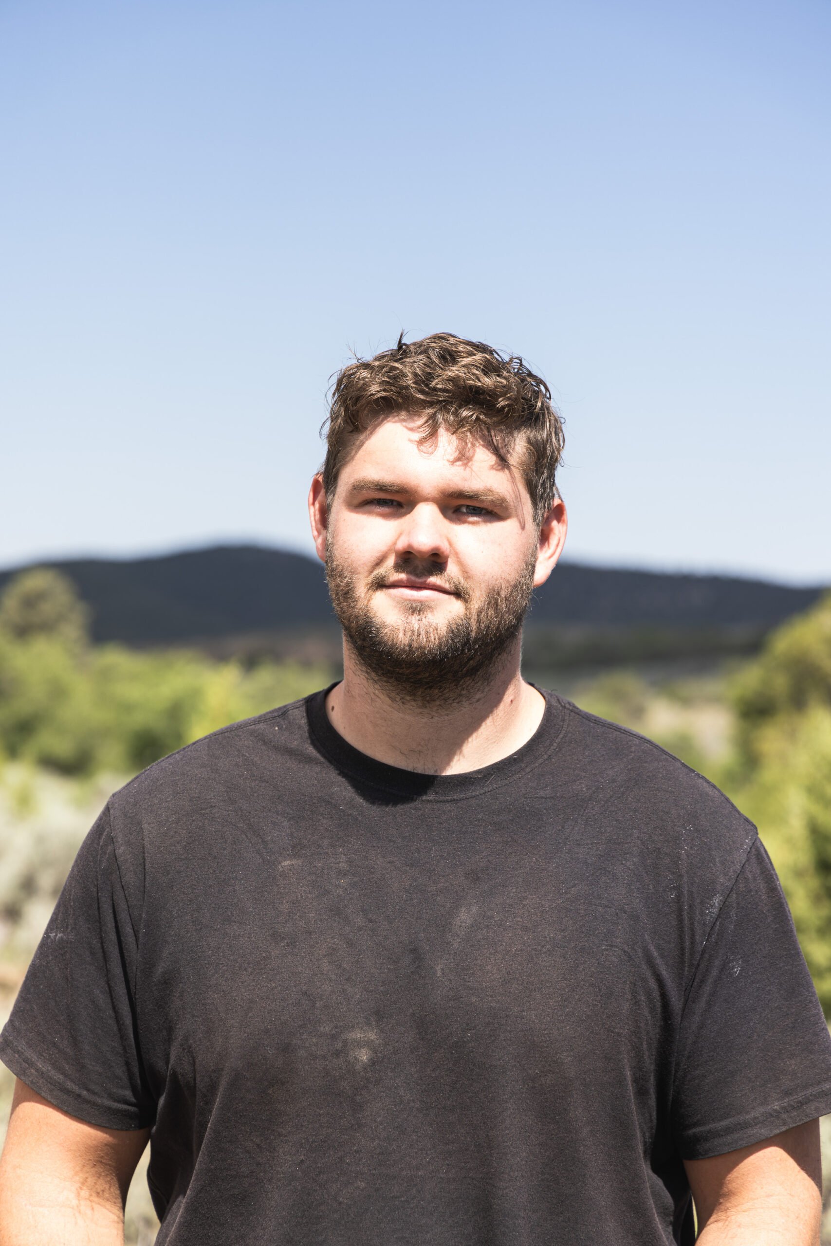 A young man with brown hair and a beard standing outdoors on a sunny day with a clear sky, green trees, and hills in the background.