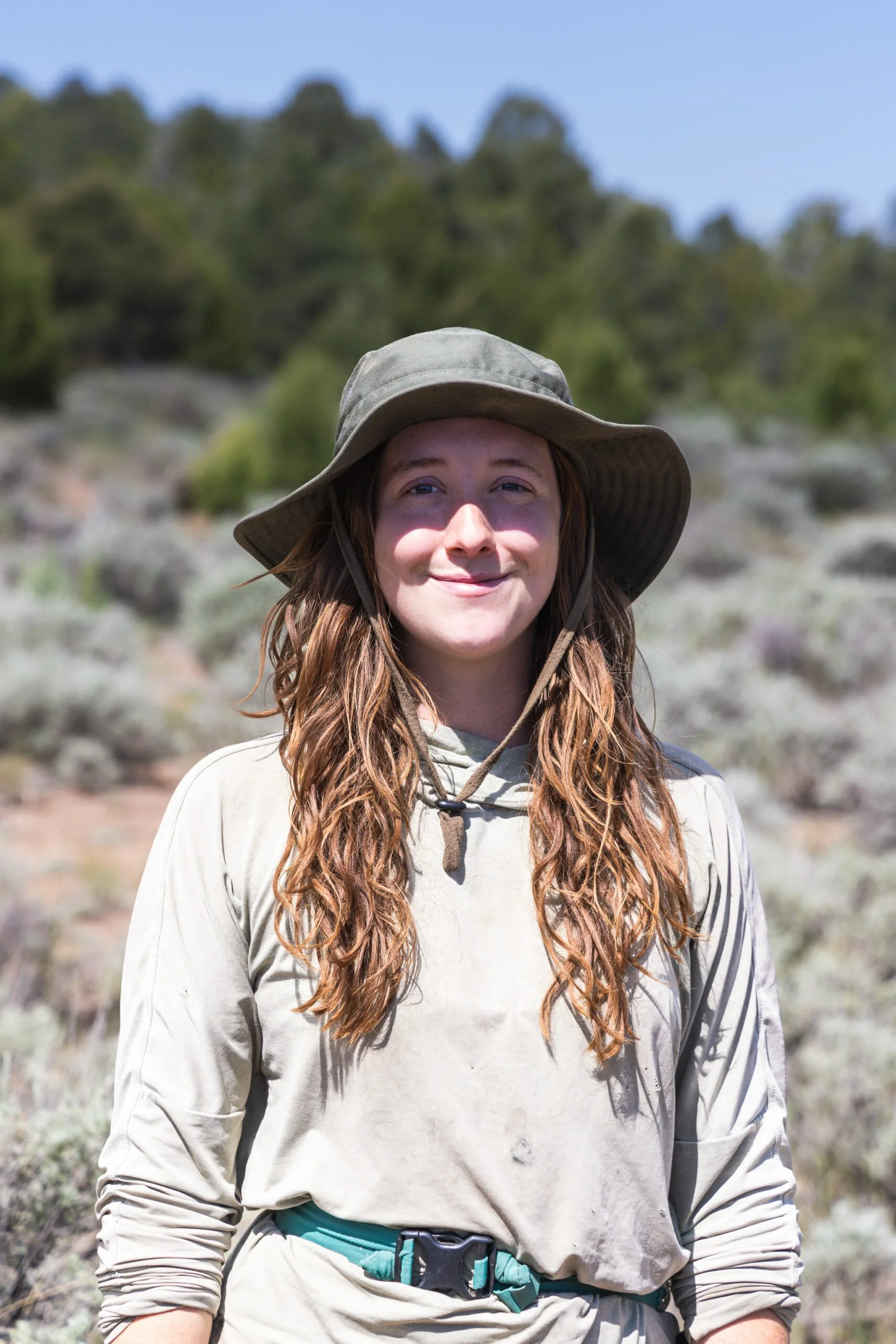 A young woman with long, curly red hair wearing a wide-brimmed hat and outdoor hiking clothing, standing in a natural landscape with bushes and trees in the background.