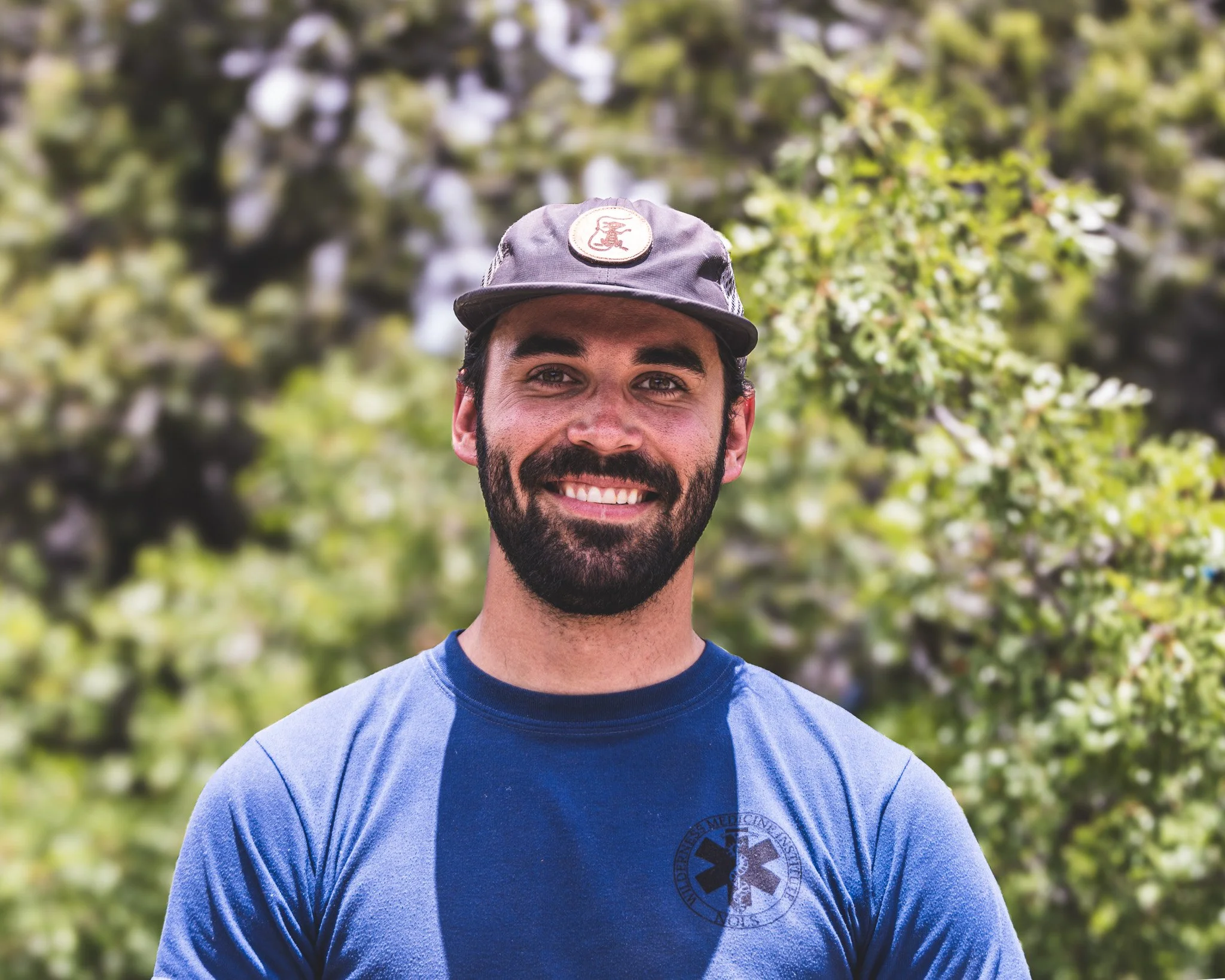 Smiling man with a beard wearing a gray cap and blue shirt stands outdoors in front of green trees.
