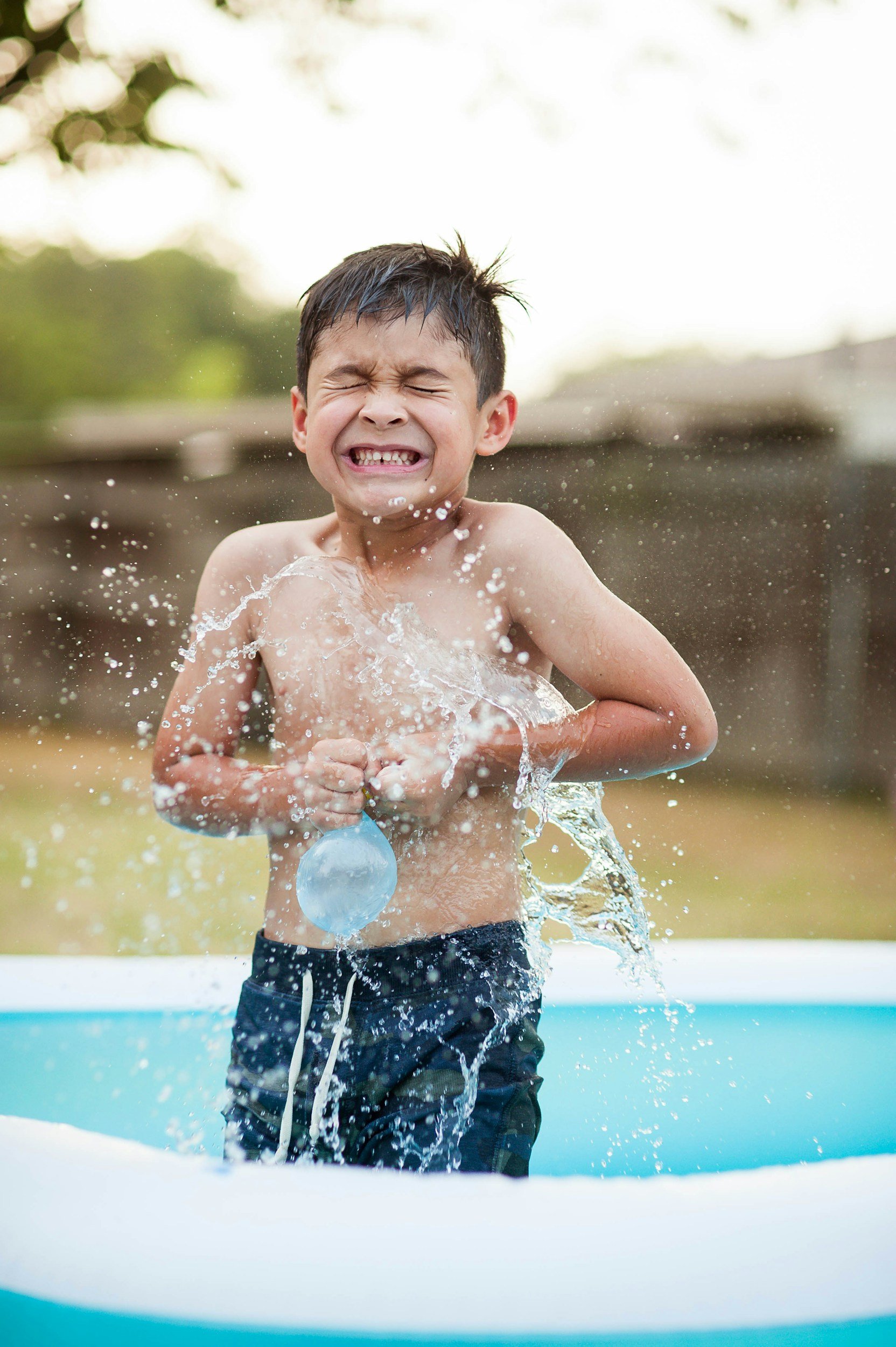 A young boy playing with water in an inflatable pool, splashing and laughing outdoors.