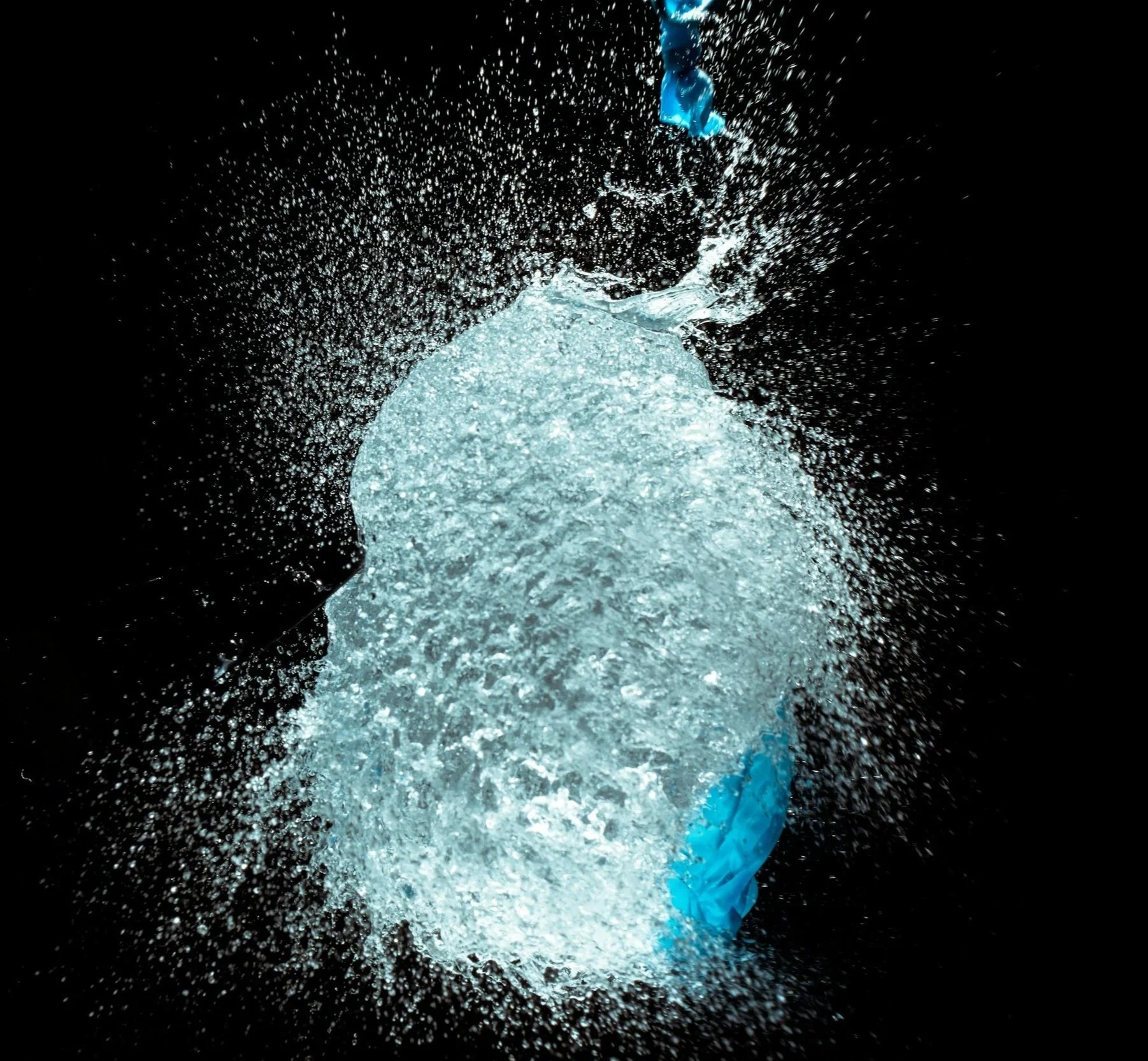 A splash of a water balloon captured mid-air against a black background, with some blue paper or plastic pieces visible at the top and bottom.