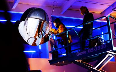 Welders on a lift working on a large sculpture of a person's face, illuminated by colorful neon lights