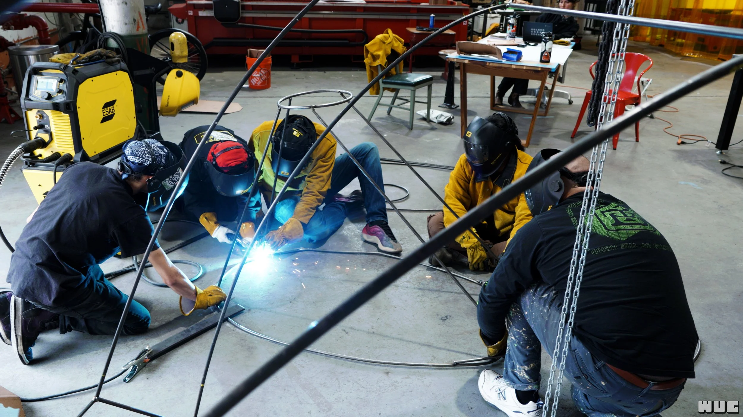 Group of five people welding a metal structure in a workshop, surrounded by tools and equipment.