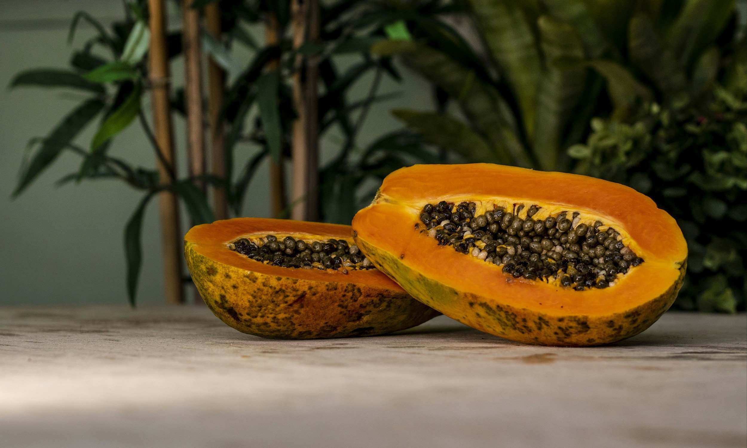 A halved papaya fruit with orange flesh and black seeds resting on a wooden surface, with green leafy plants in the background.