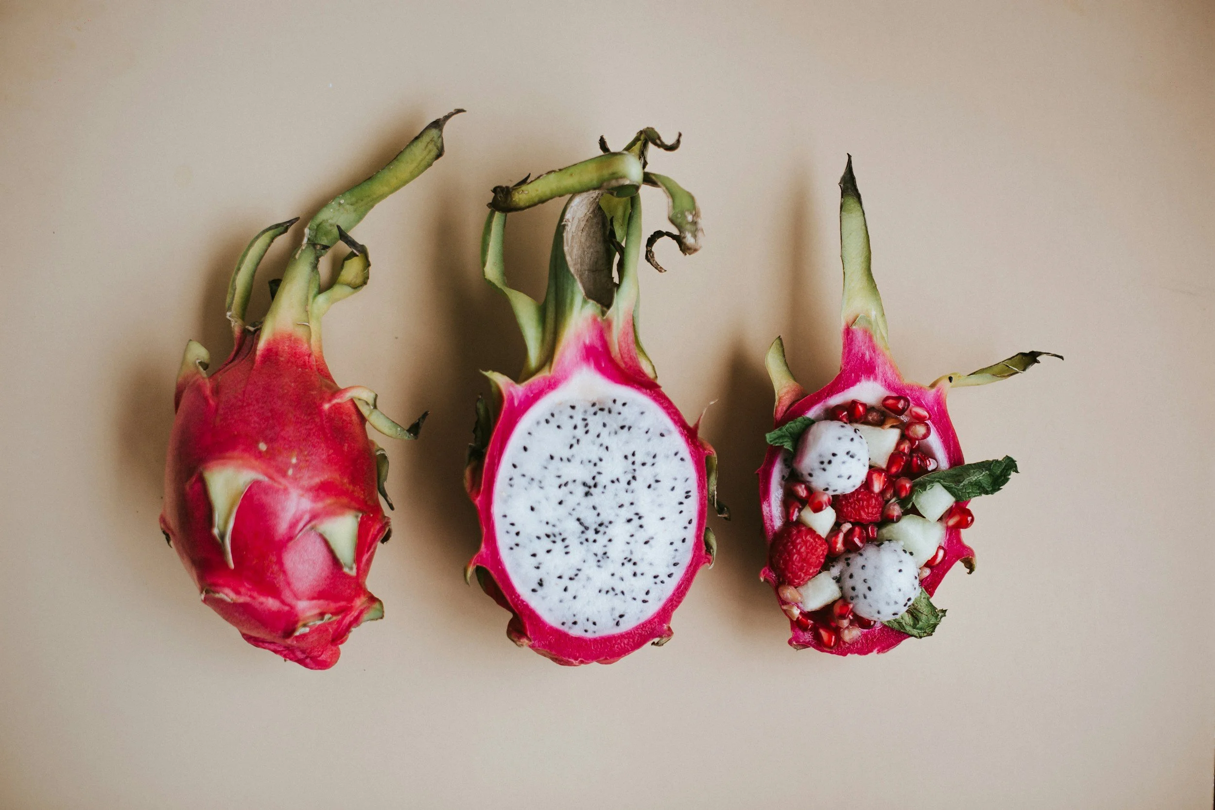 Three dragon fruits, one sliced open showing white flesh with black seeds, and the other two halved with red and white flesh and surrounded by various small fruits.
