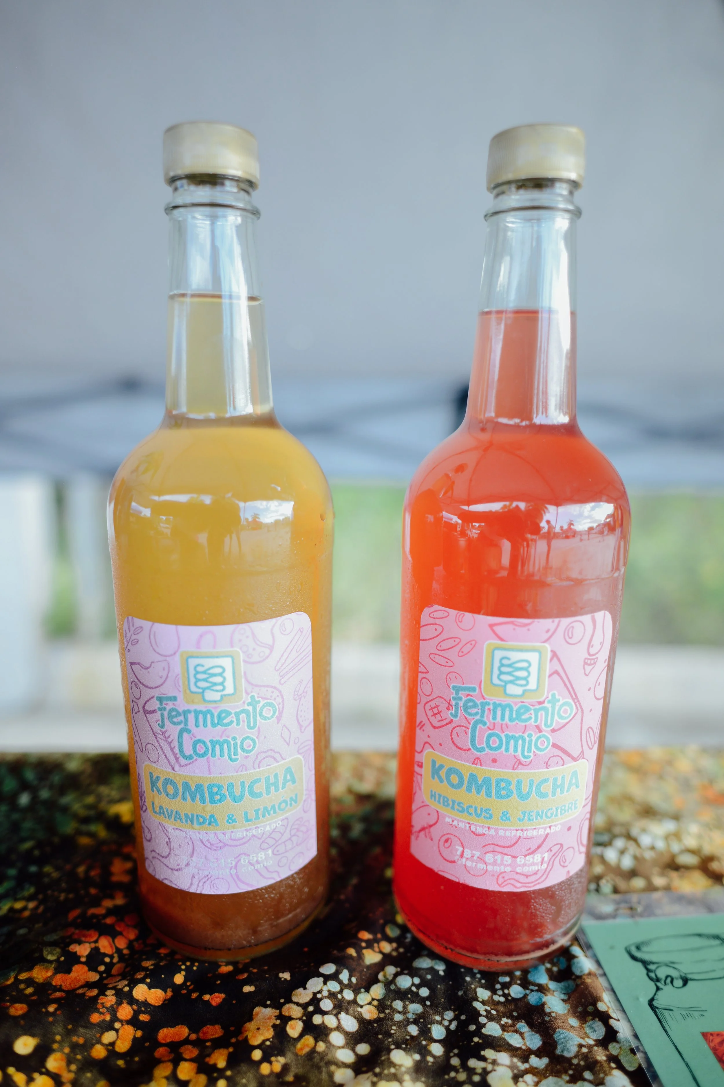 Two bottles of kombucha placed on a surface with a colorful, speckled pattern. The labels on the bottles indicate different flavors, with one being lavender lemon and the other hibiscus ginger. The background is blurred, with a tent or canopy visible.