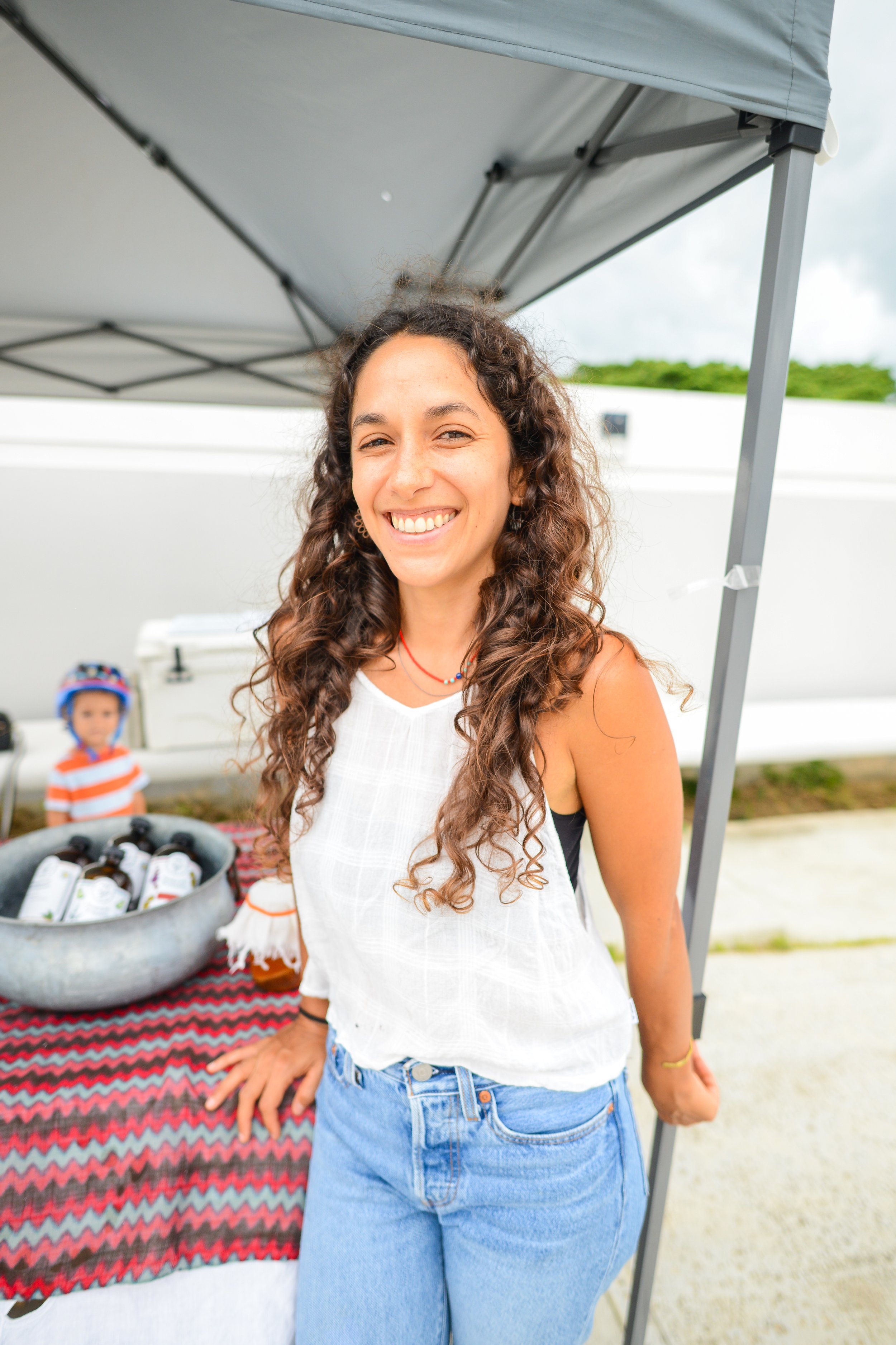 A woman with long curly brown hair smiling, wearing a white sleeveless top and blue jeans, standing under a canopy at an outdoor event. In the background, a young child is visible behind a table with a red, black, and white tablecloth, and various bottles. The setting appears to be a daytime outdoor gathering.