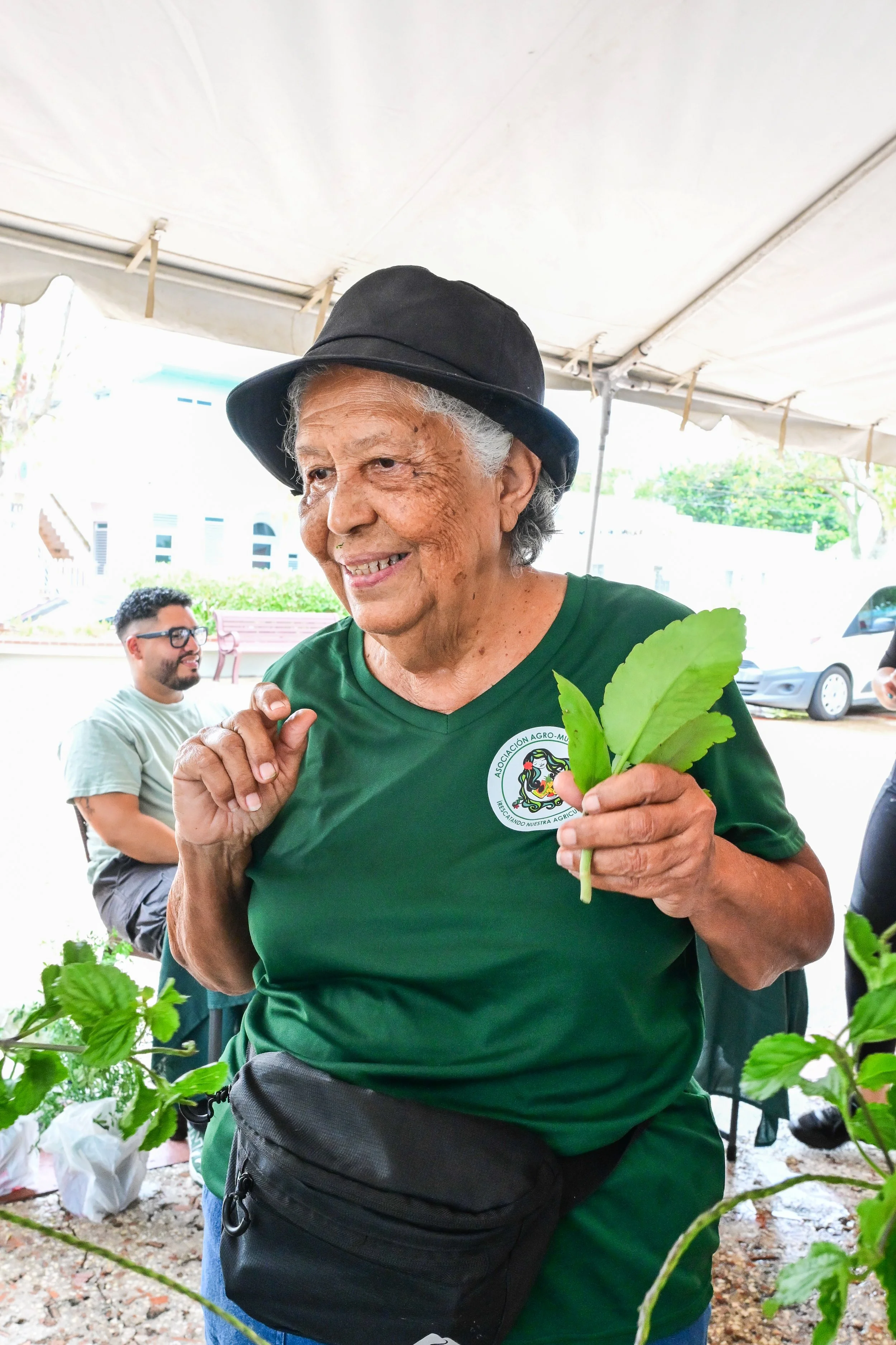 A smiling elderly woman with gray hair, wearing a black hat and green T-shirt, holding a green plant leaf at an outdoor gathering under a tent.