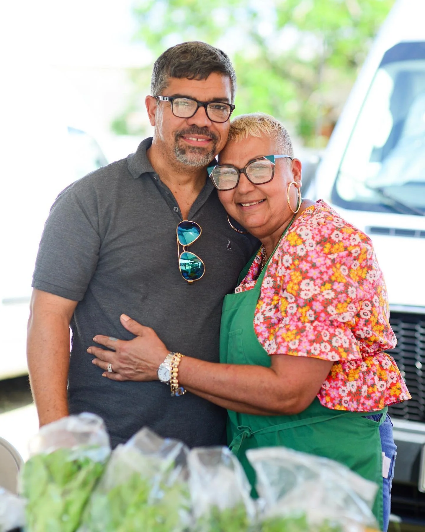 A man and woman are standing close together, smiling and embracing each other. They are both wearing glasses. The man has short dark hair and a beard, wearing a gray polo shirt. The woman has short blonde hair, wearing a colorful floral blouse and a green apron. The background shows a garage or workshop setting with a vehicle and some greenery outside. The foreground has plastic bags or containers.