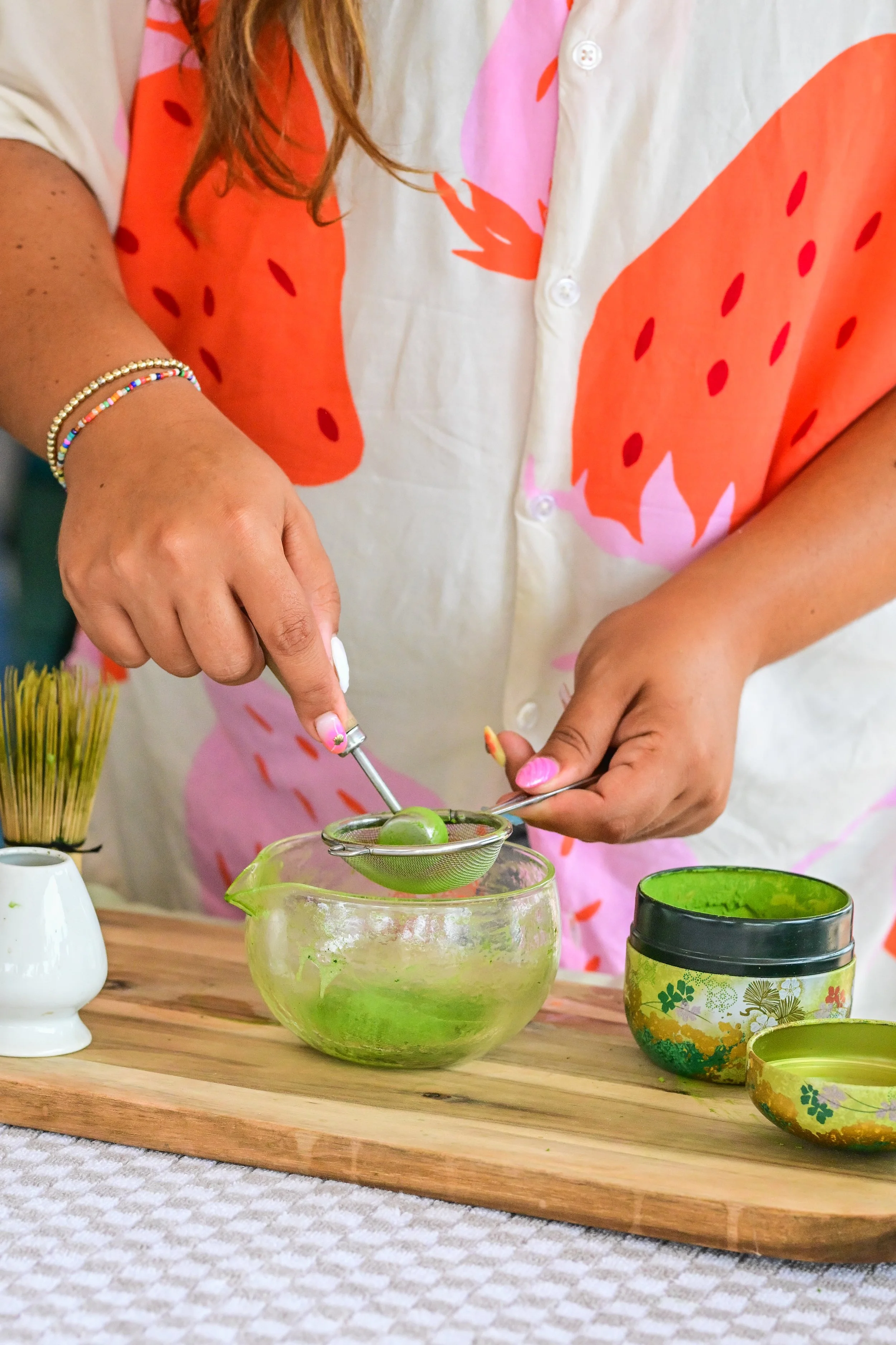 A person wearing a white shirt with a pink and orange watermelon design is preparing matcha tea using a sifter and a bowl of green tea. Various cups and a jar of matcha powder are on a wooden tray.