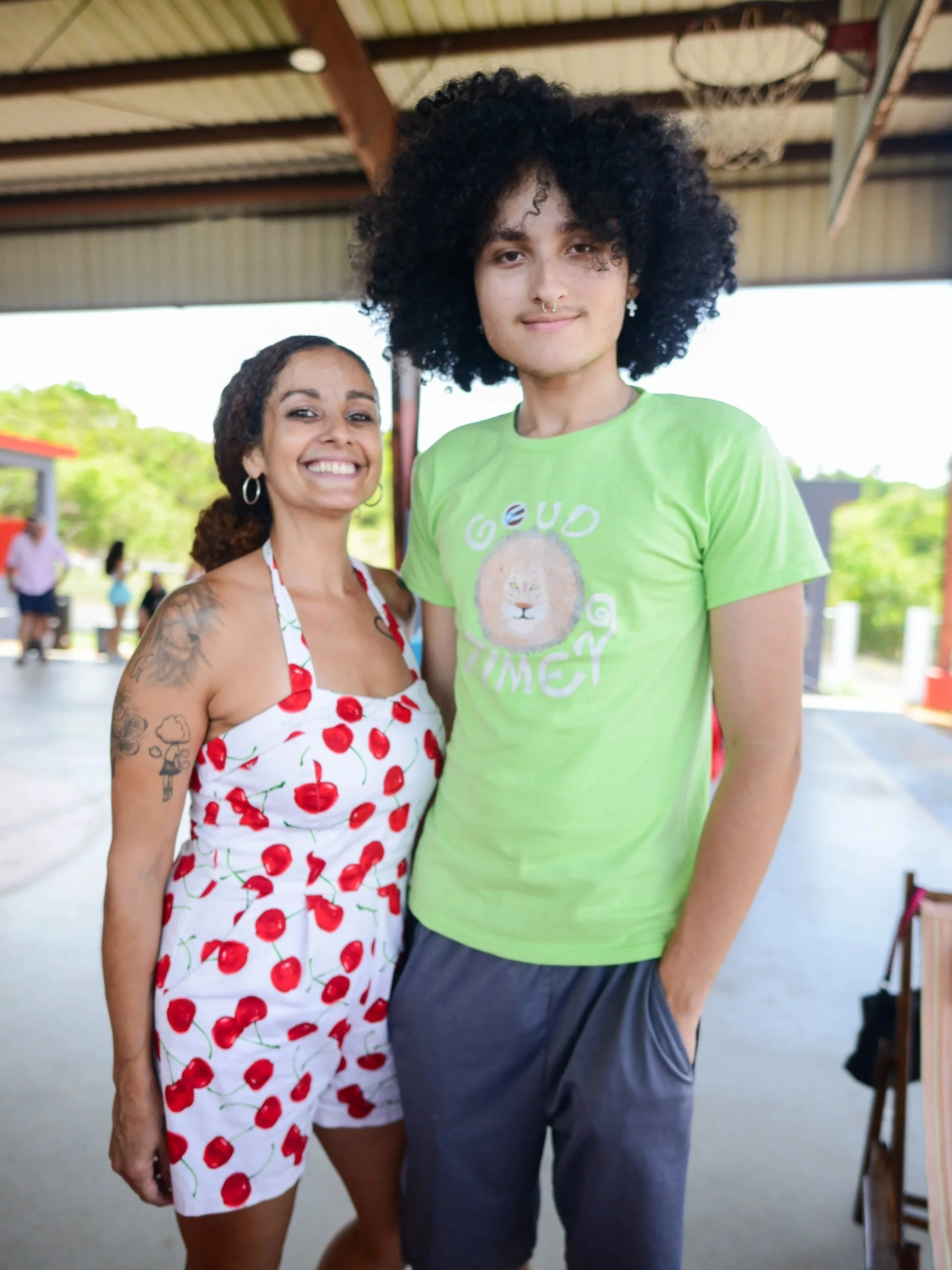 Two smiling women standing under a metal roof at an outdoor event, one wearing a dress with cherry print and the other in a bright green T-shirt.