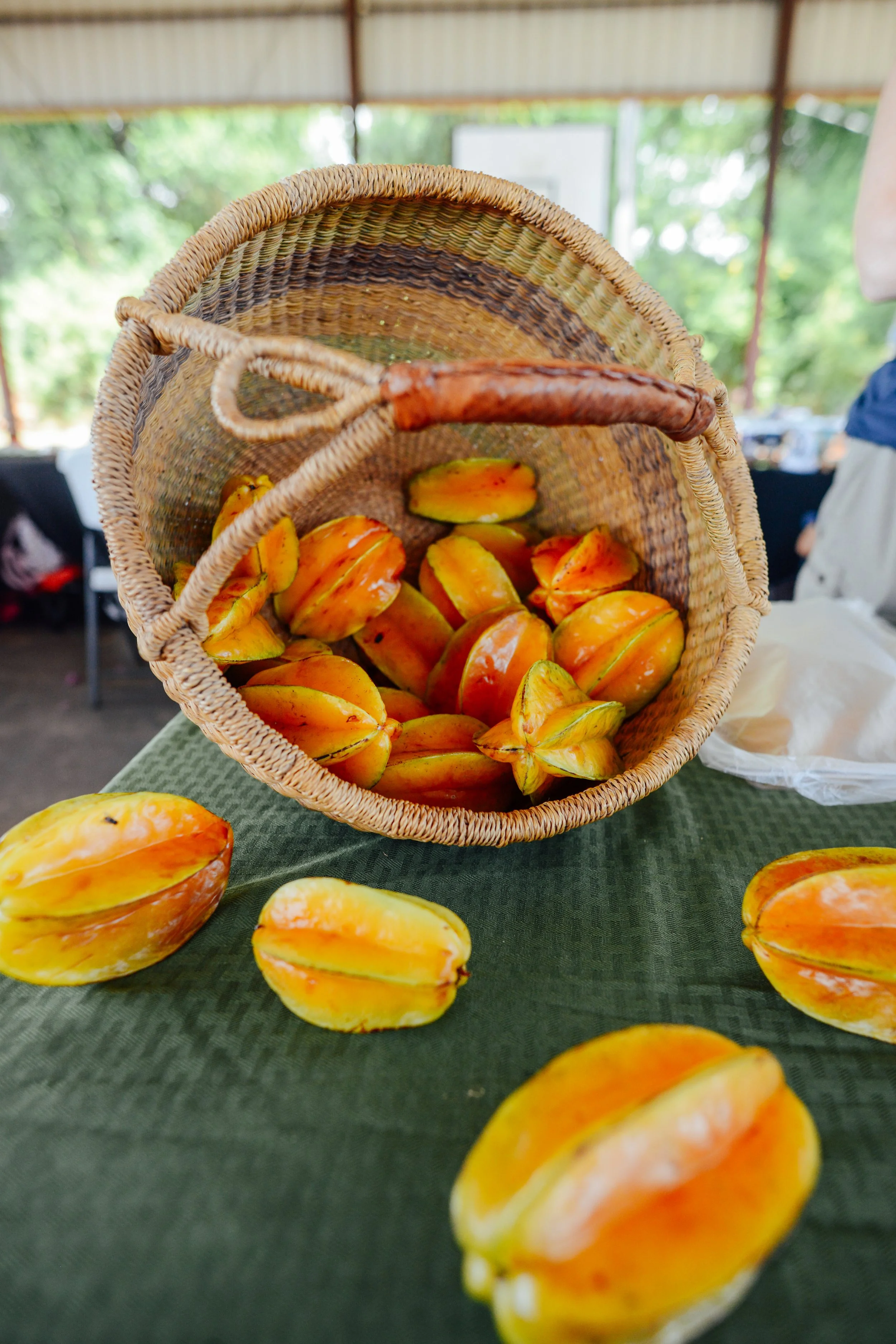 A basket of starfruit slices on a green tablecloth, with some slices also scattered in front of the basket, set outdoors under a covered area with trees in the background.