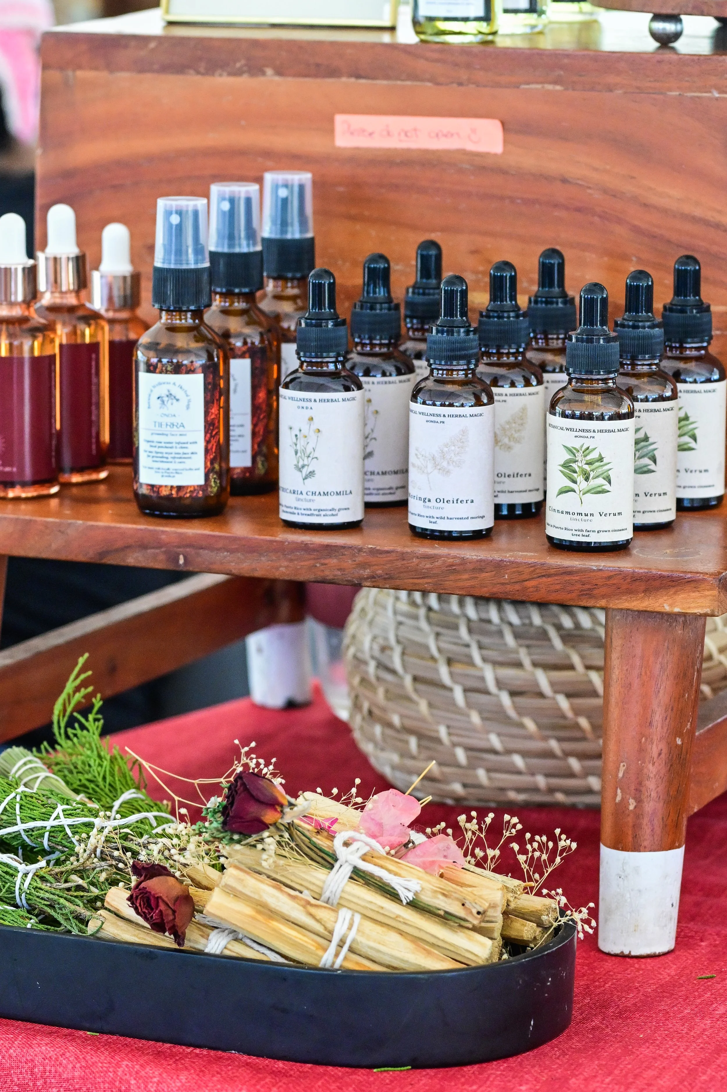A display of essential oils in amber glass bottles with dropper caps, arranged on a wooden shelf. The bottles have botanical labels featuring illustrations of herbs and plants. In the foreground, there is a black tray filled with dried flowers and herbs, and below it, a woven basket with a plant. The scene has a warm, cozy ambiance.